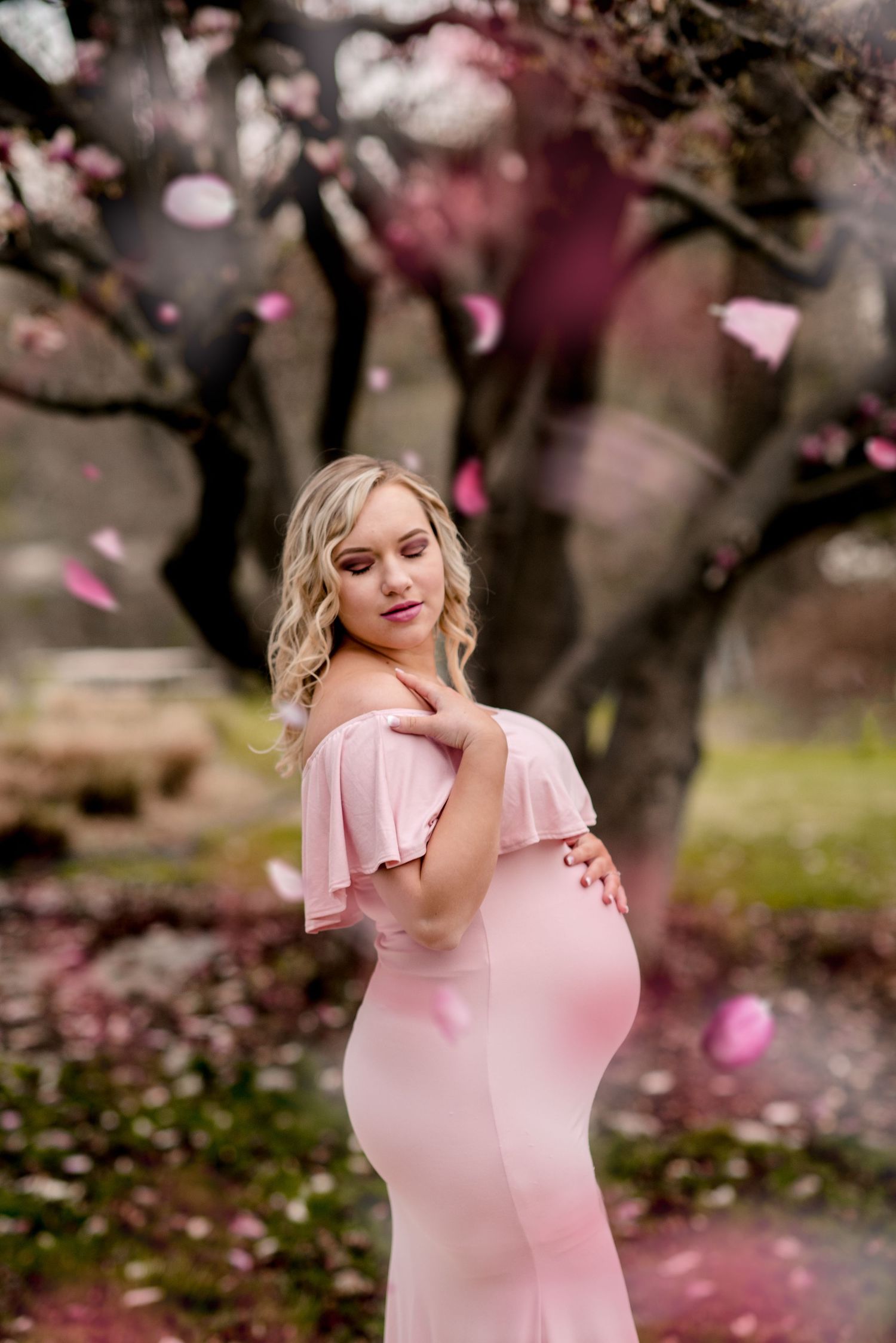 Woman in pink dress poses gracefully among falling cherry blossom petals in spring garden.