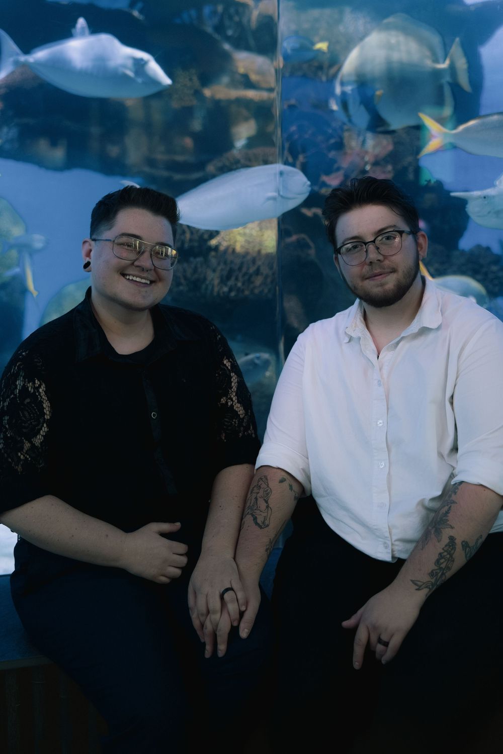 Two people sit together in front of a large aquarium tank filled with swimming fish and blue lighting.