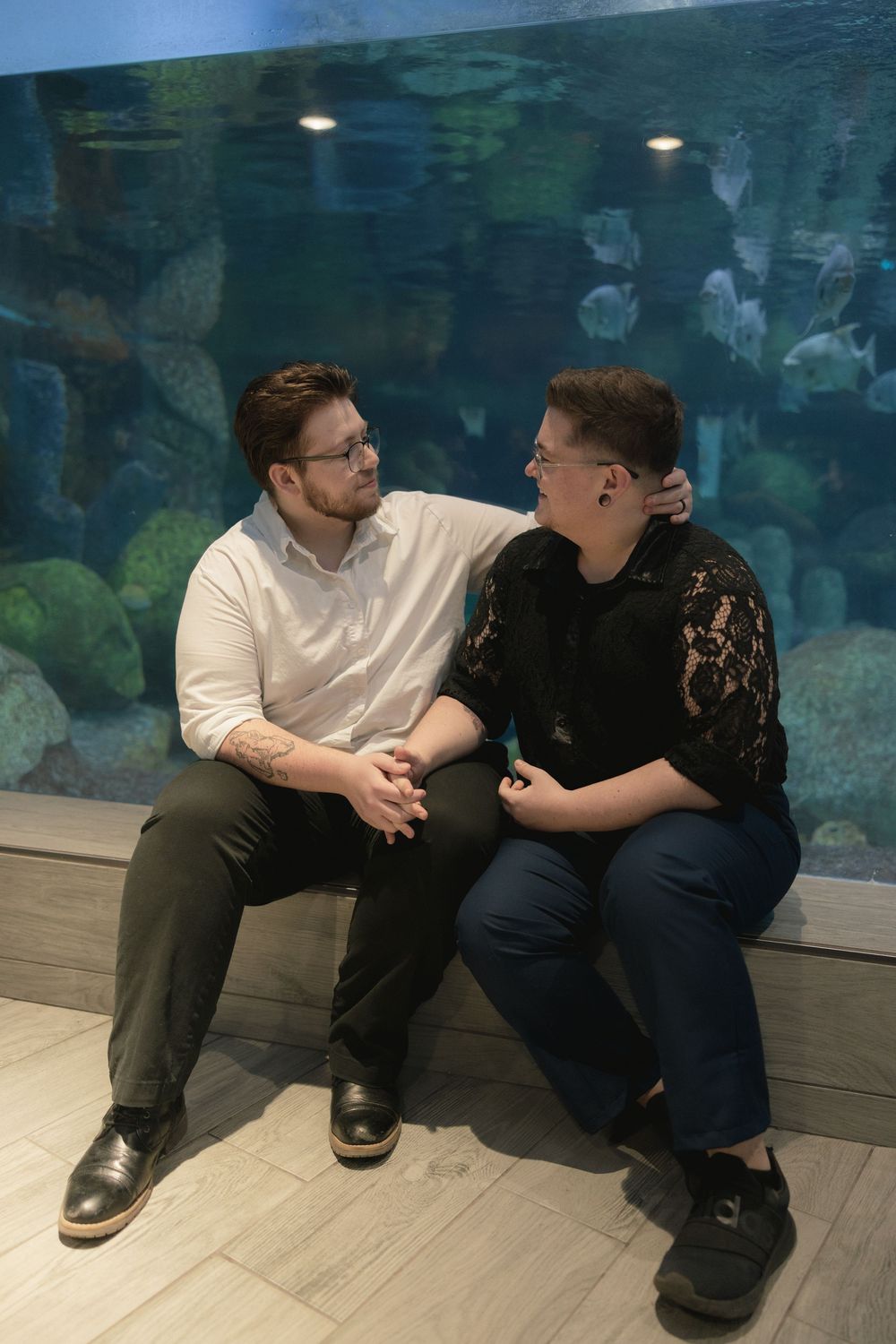 A couple sits together on a bench in front of a large aquarium display with blue-green lighting.