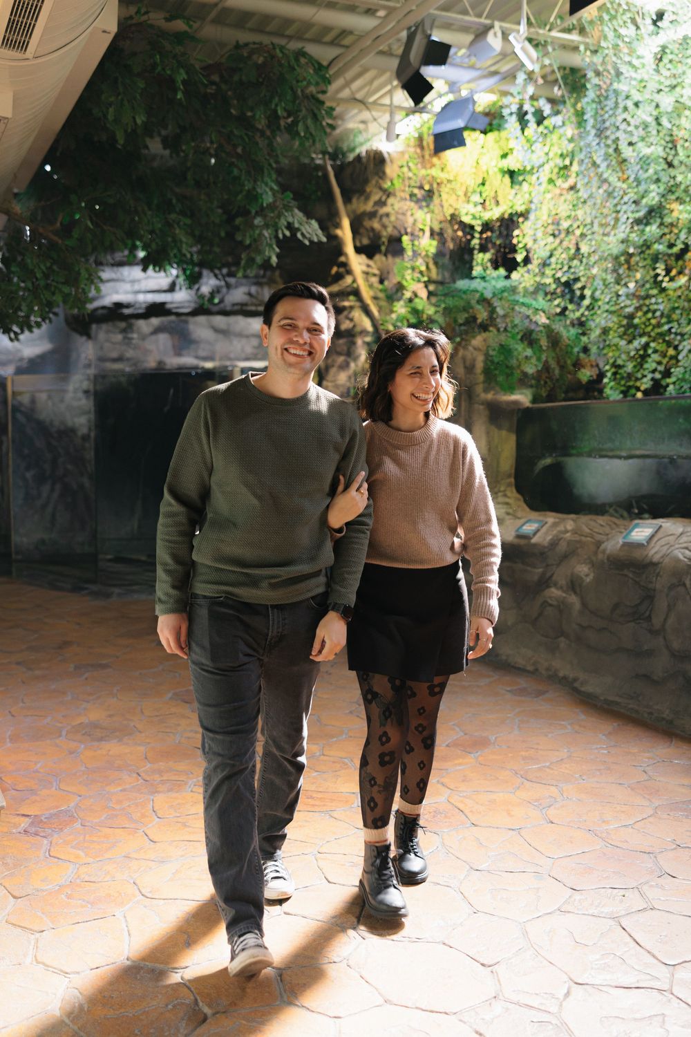 Two people walk together outdoors on a terracotta tiled patio with greenery and natural lighting in the background.
