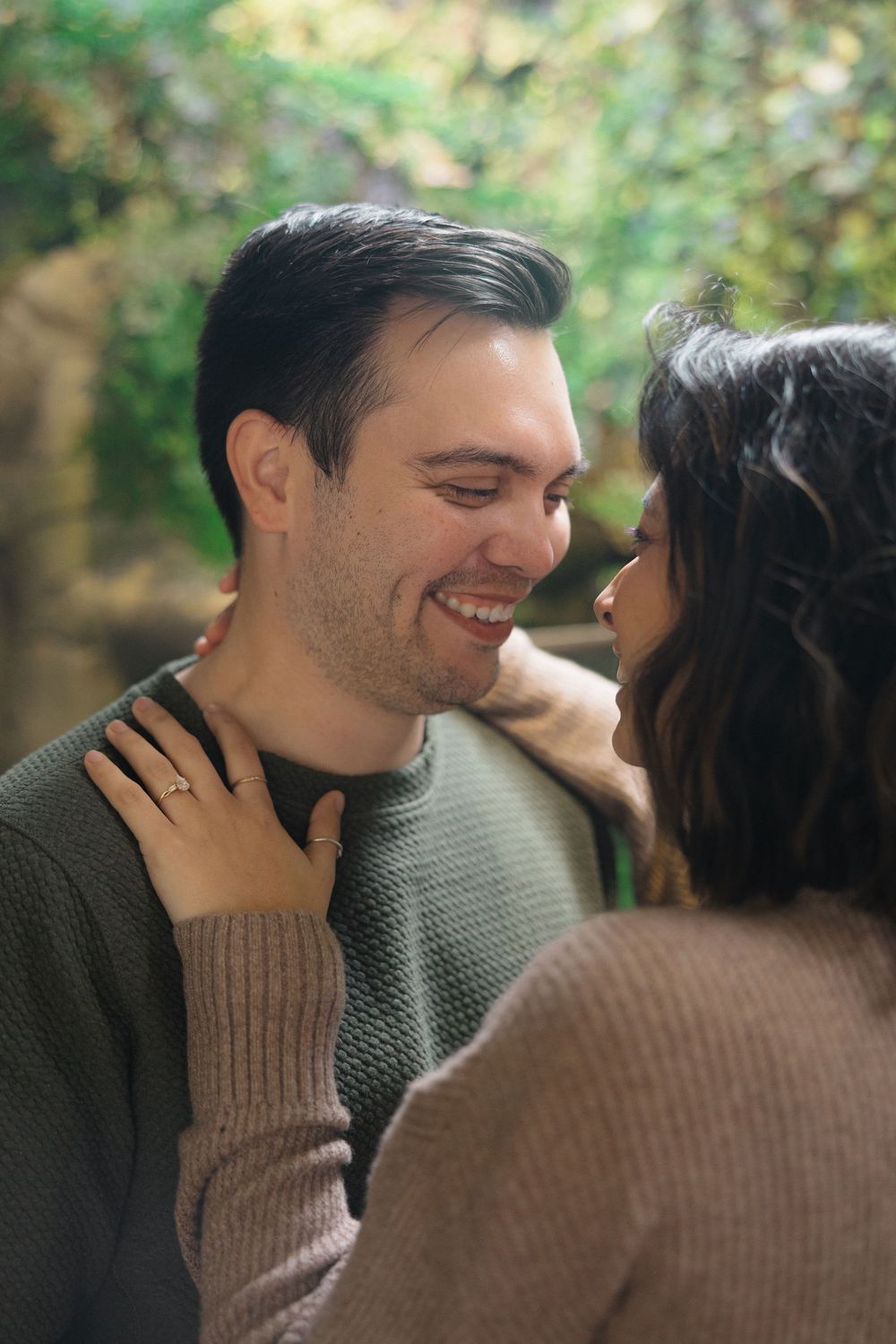 A romantic couple in cozy sweaters share an intimate moment outdoors with natural greenery in the background.