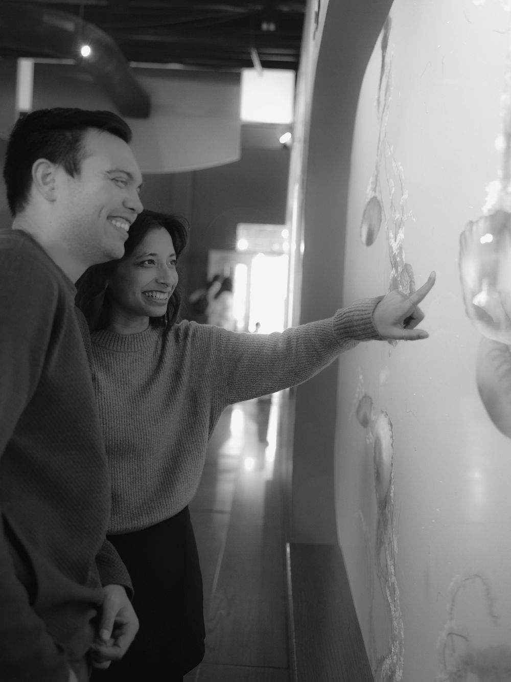 Two people in casual attire smile and point at something on a wall in what appears to be an indoor gallery space.