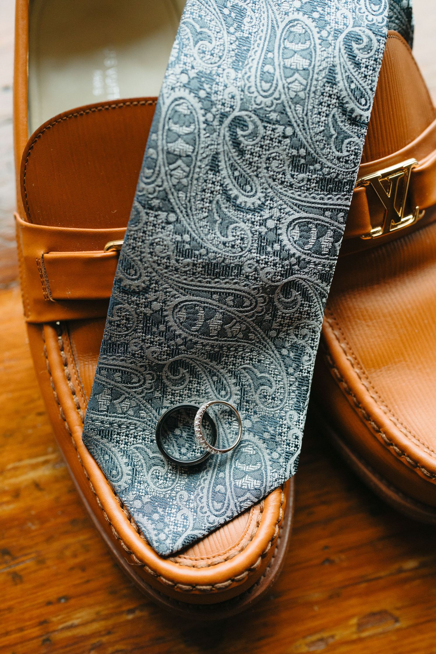 Blue paisley tie rests against brown leather dress shoes on wooden floor.