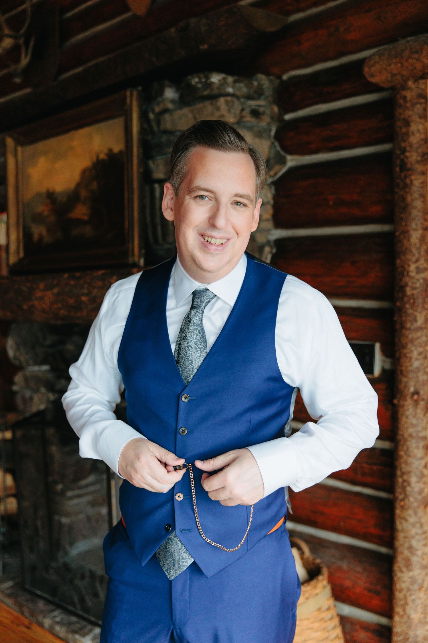 Person in formal attire wearing a blue vest and tie adjusts their pocket watch chain in a rustic wooden interior setting.