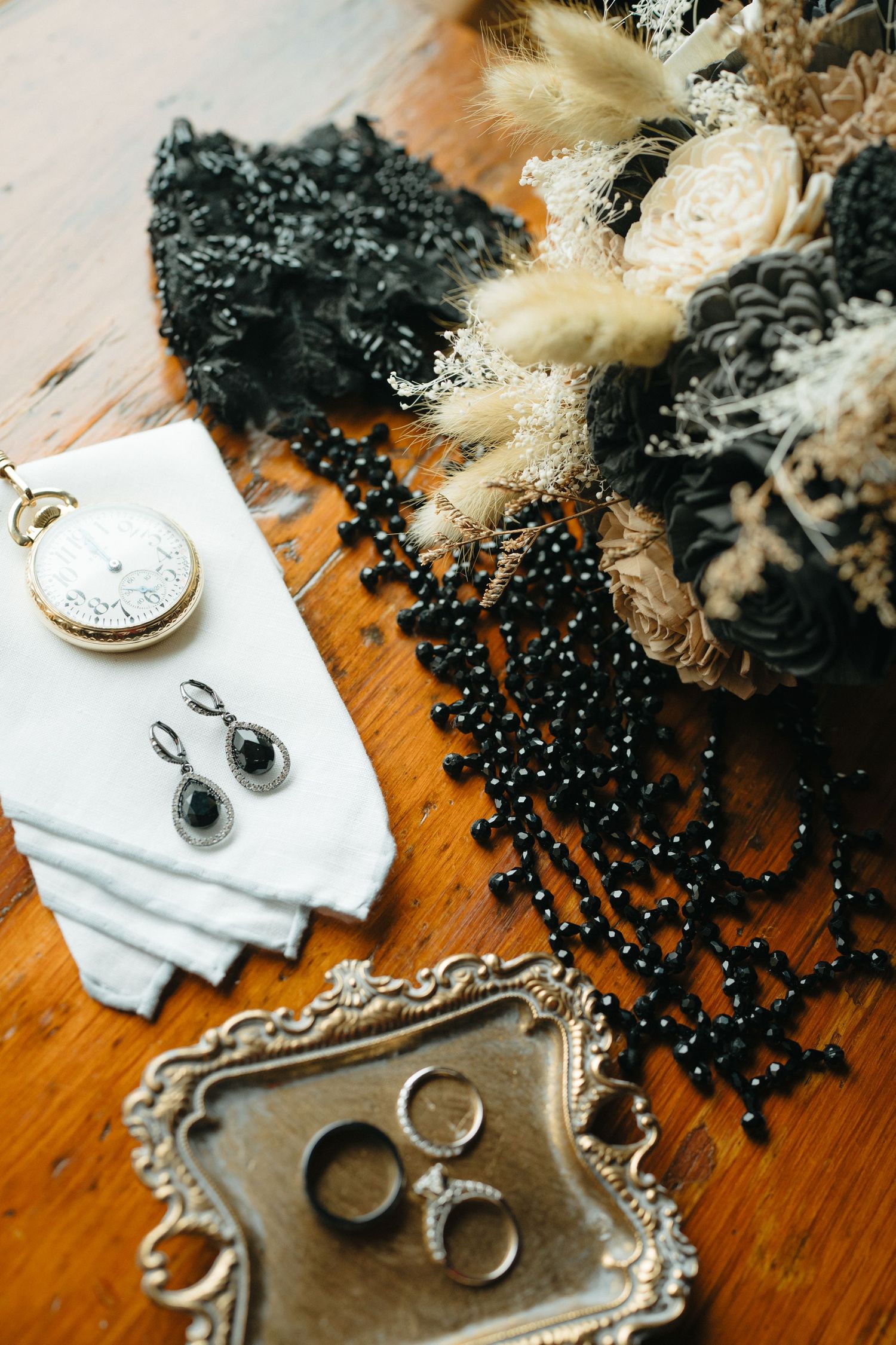 Vintage wedding accessories including pocket watch, black beaded necklace, rings on ornate silver tray, and earrings on white fabric.