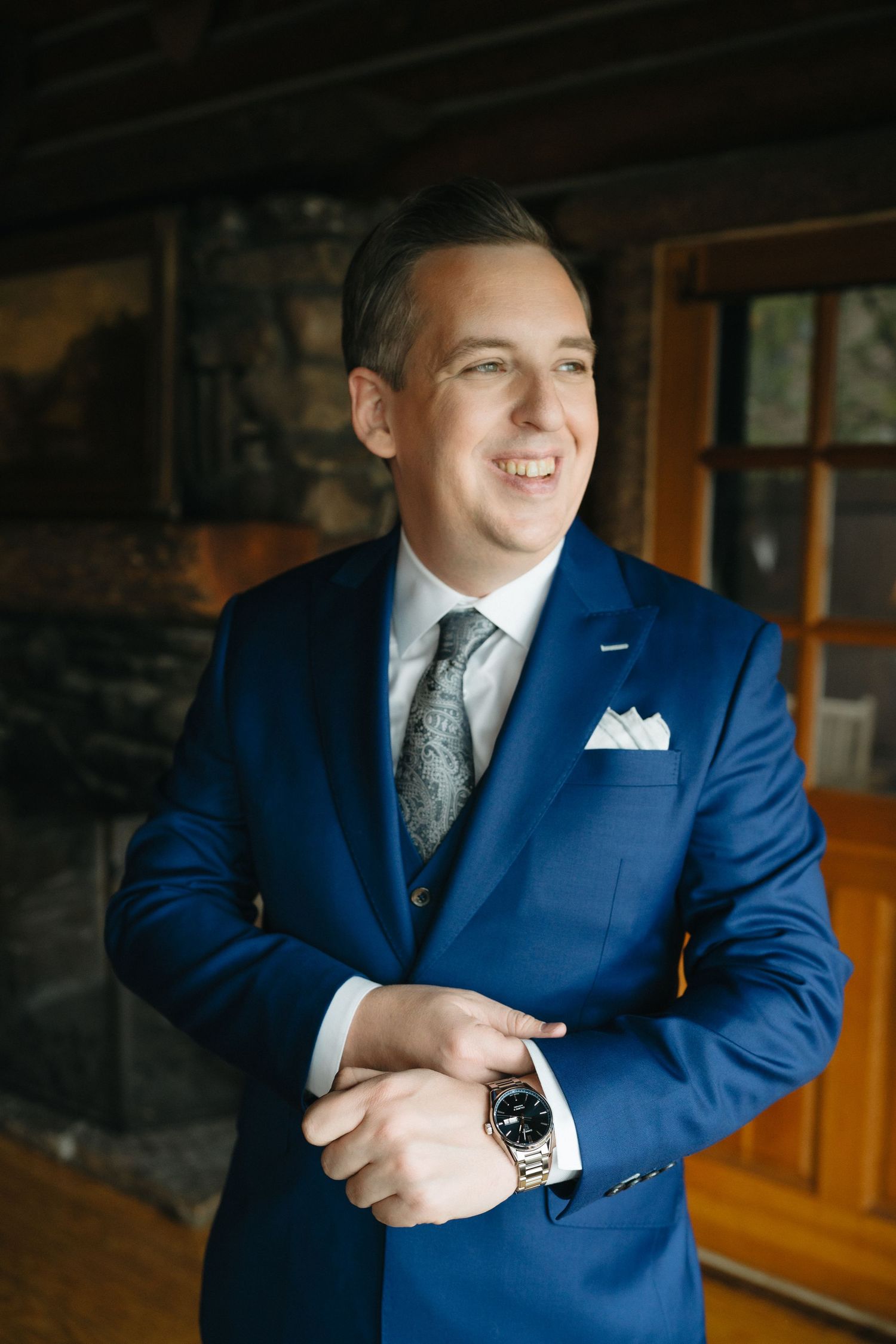 A groom in a tailored navy blue suit adjusts his cufflinks while standing in a rustic indoor setting.