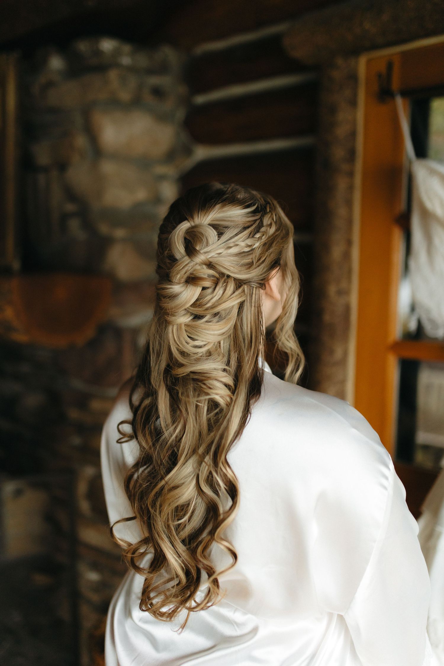 Back view of a person in a white satin robe with styled blonde wavy hair in a rustic room.