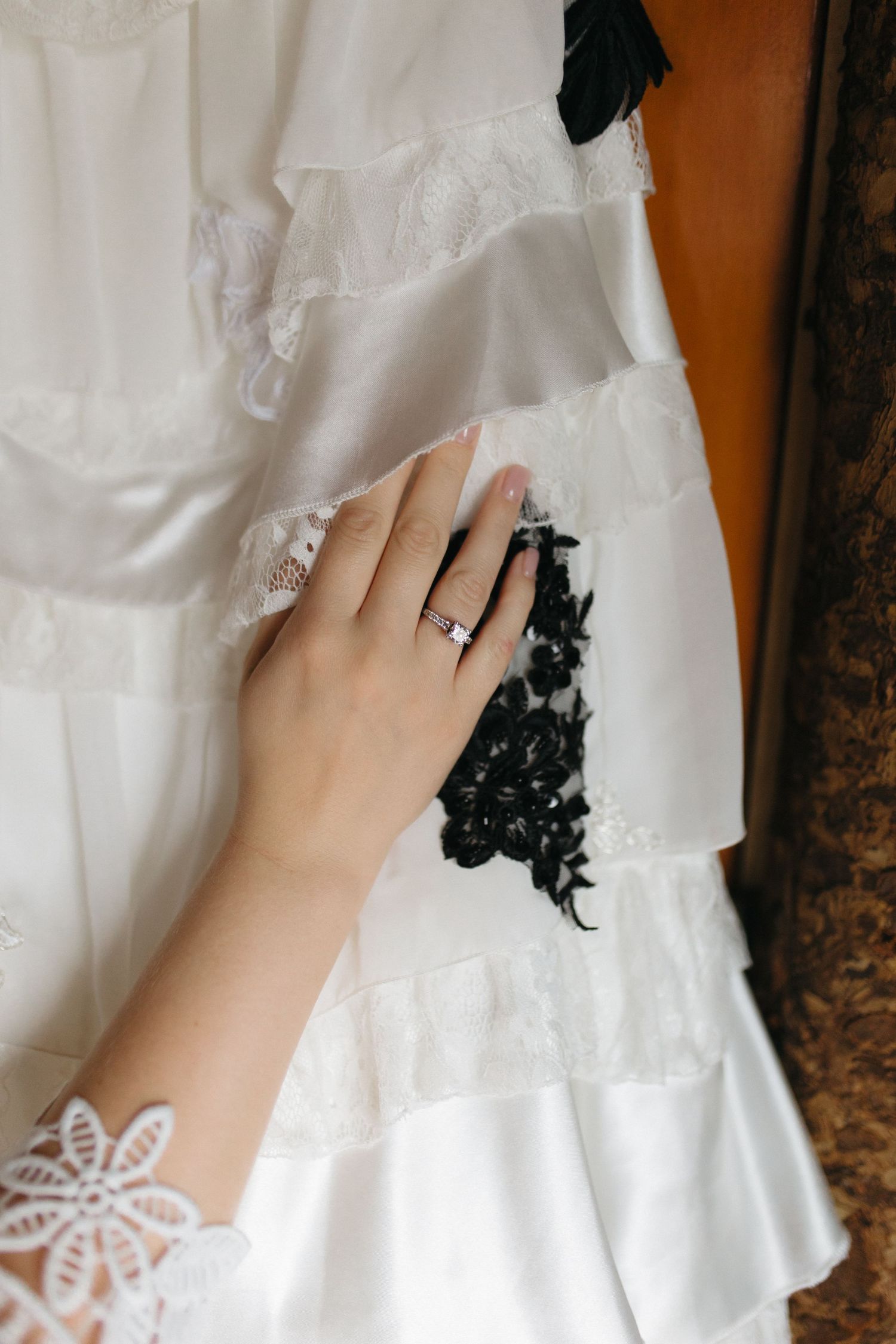 Close-up view of delicate white lace details and a sparkling engagement ring on a hand against a dark background.