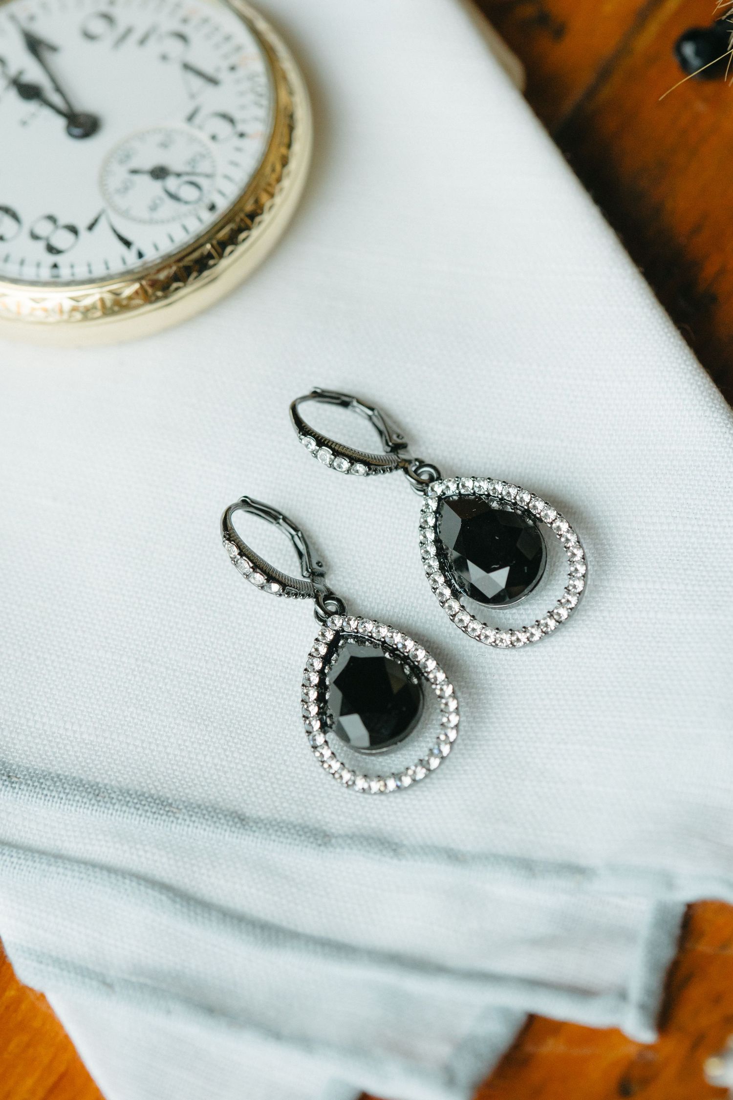 Elegant black teardrop earrings with crystal accents displayed on white fabric next to vintage timepiece.