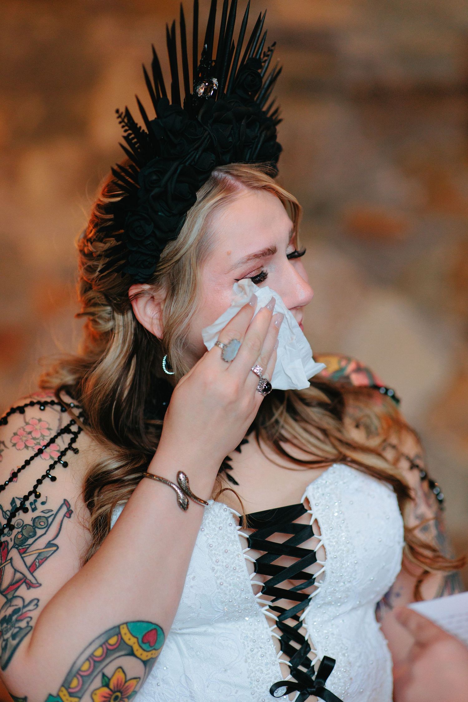 A bride wearing a black spiked crown and white dress wipes tears with a tissue.