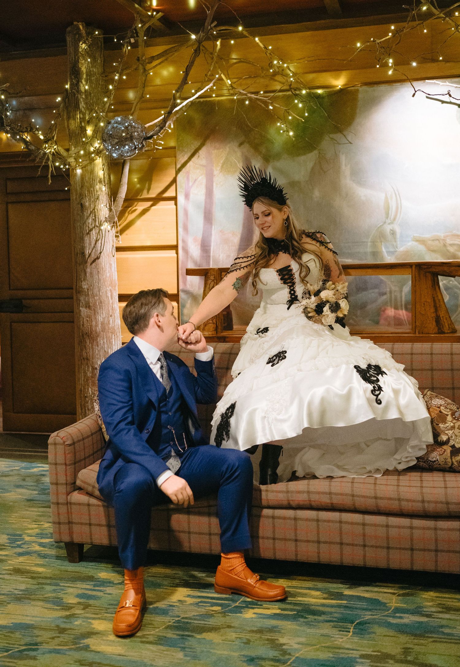 Wedding couple sits on brown couch with bride in white dress and groom in blue suit with orange shoes.