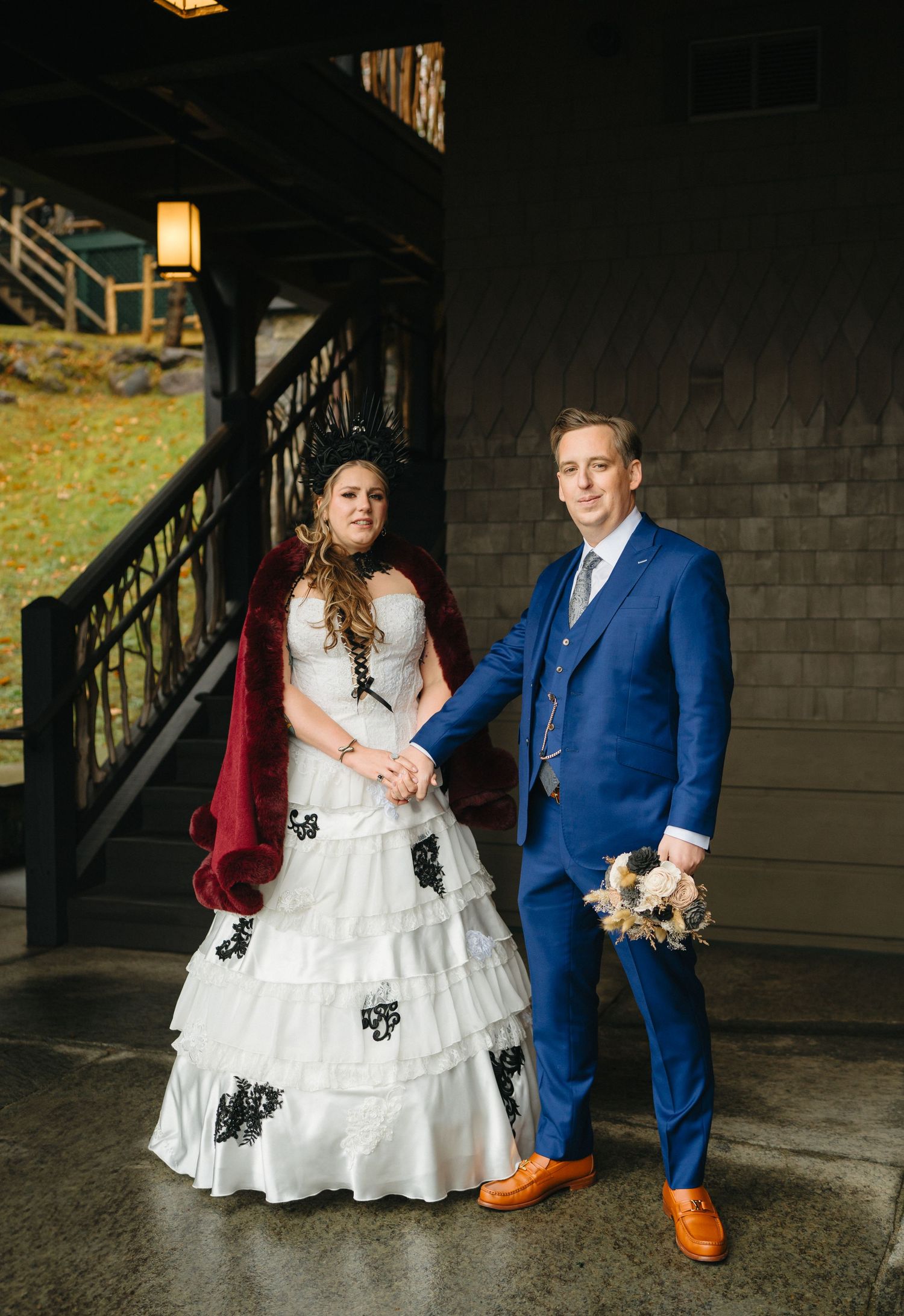 A couple in formal attire stand together under moody lighting, with the groom wearing a bright blue suit and tan shoes.
