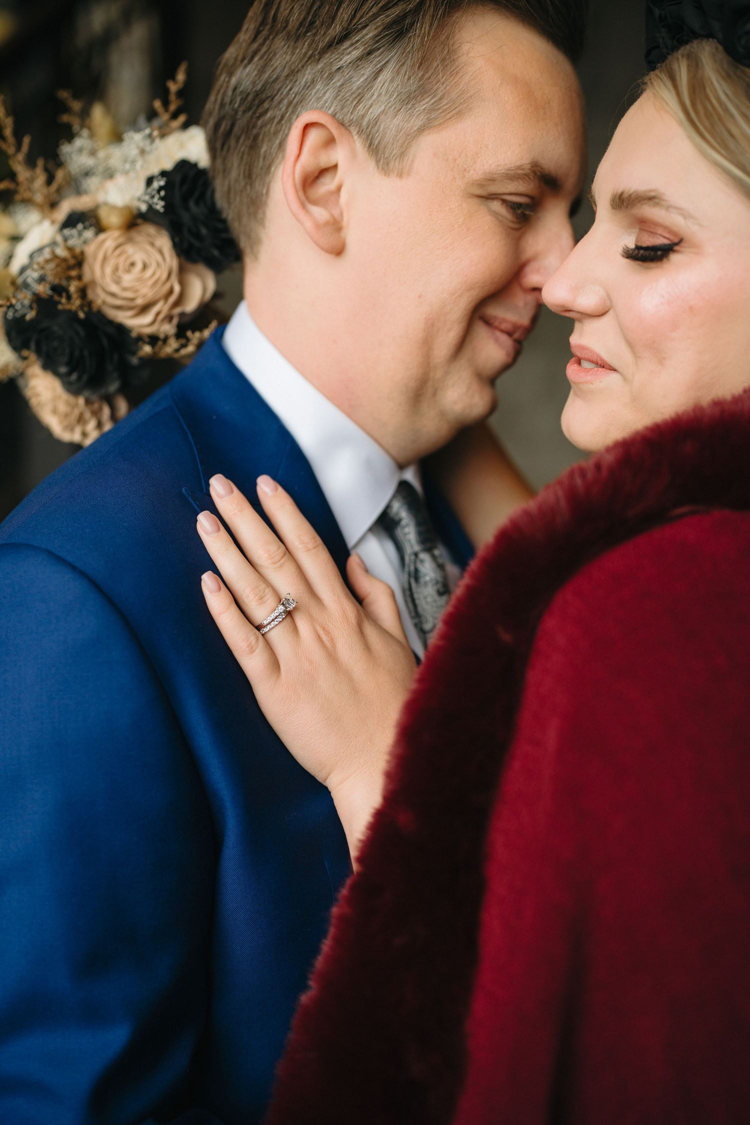 A romantic wedding portrait showing a couple in a close embrace with dried flowers and a burgundy fur wrap.