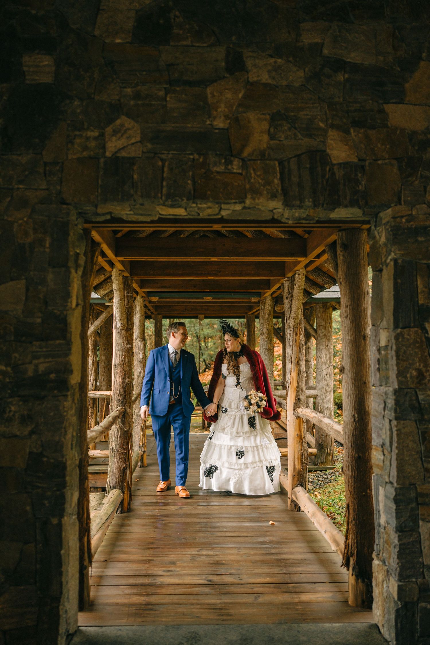 A couple in wedding attire walks through a rustic covered wooden bridge walkway.