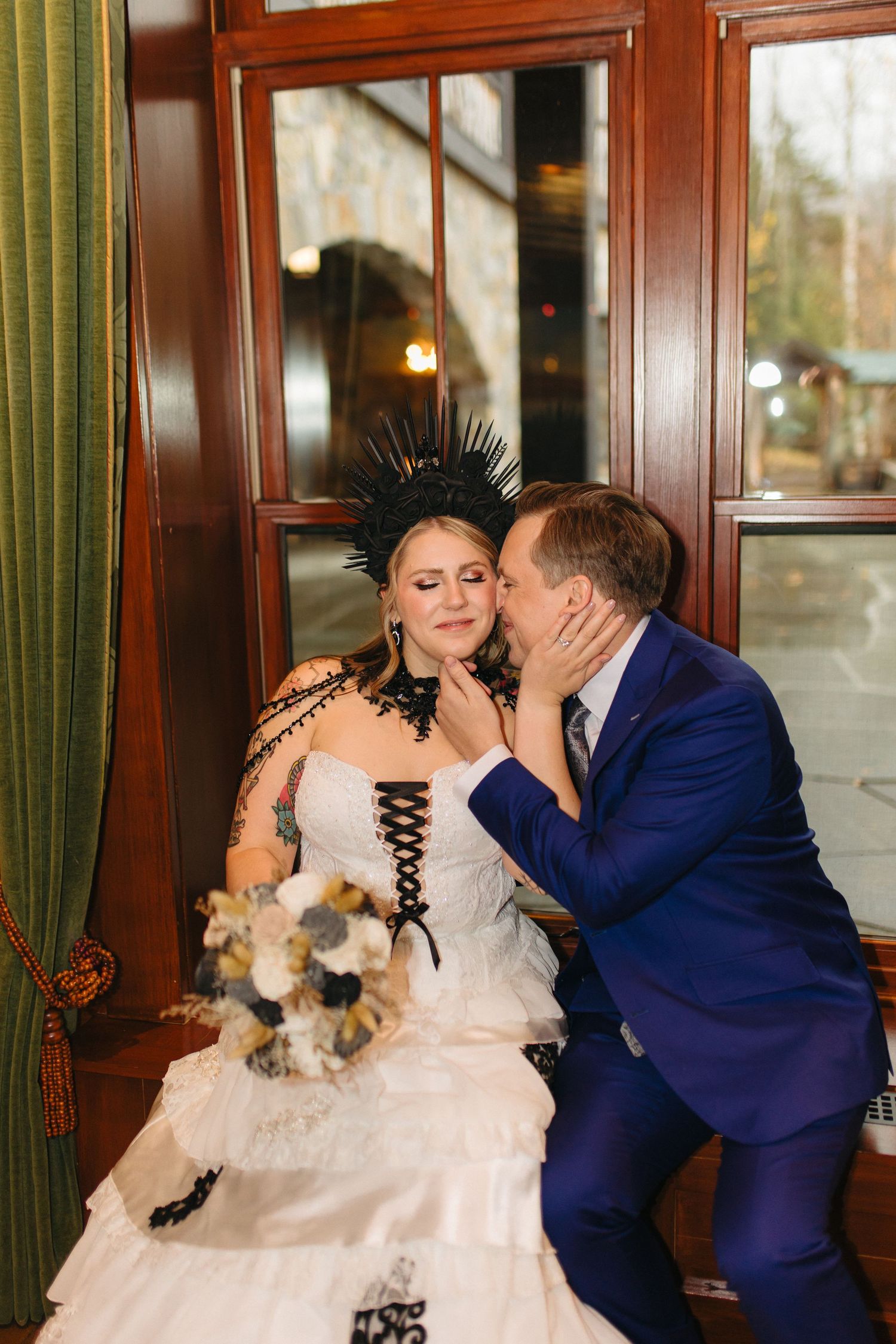 A couple shares an intimate moment in formal wedding attire with a white dress and blue suit by a window.