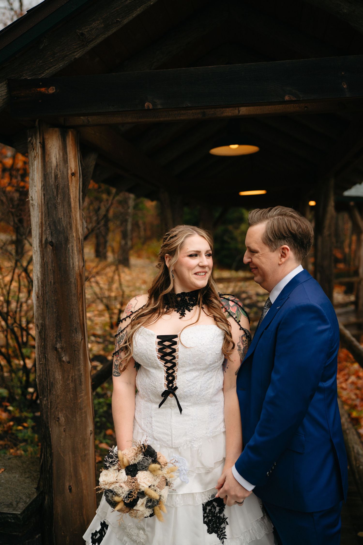 A wedding couple poses under a wooden pavilion surrounded by autumn foliage.