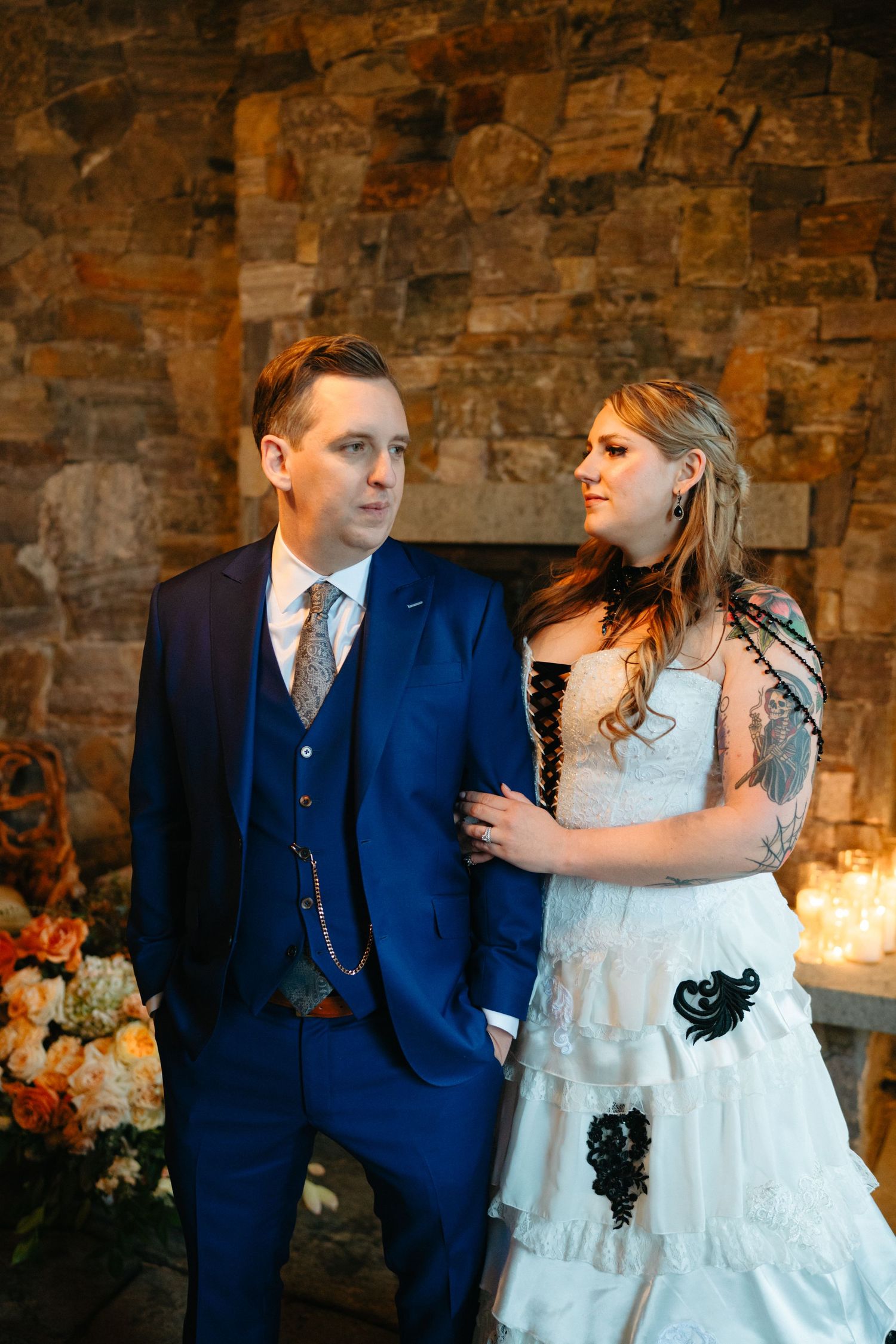 A wedding couple poses against a stone wall backdrop in elegant attire.