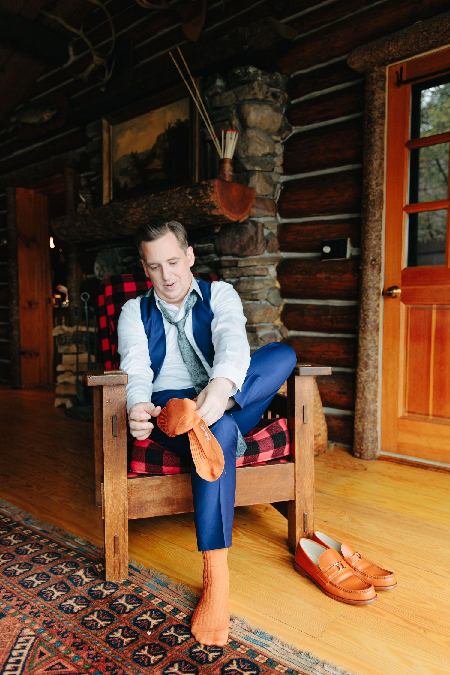 Someone puts on orange loafers while sitting in a rustic log cabin with plaid cushions and patterned rugs.