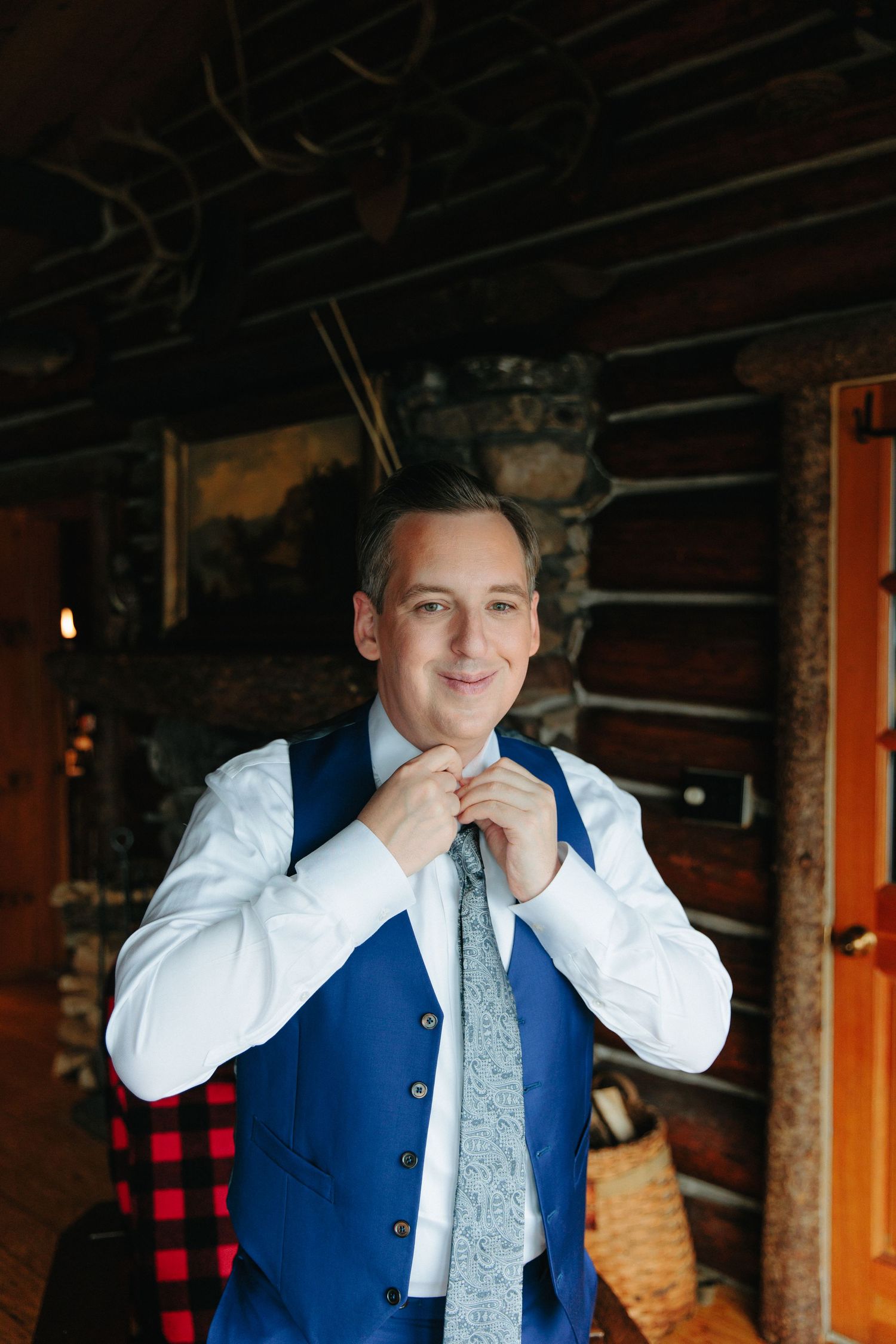 Person in blue suit vest and white shirt smiles while adjusting their tie in rustic indoor setting.