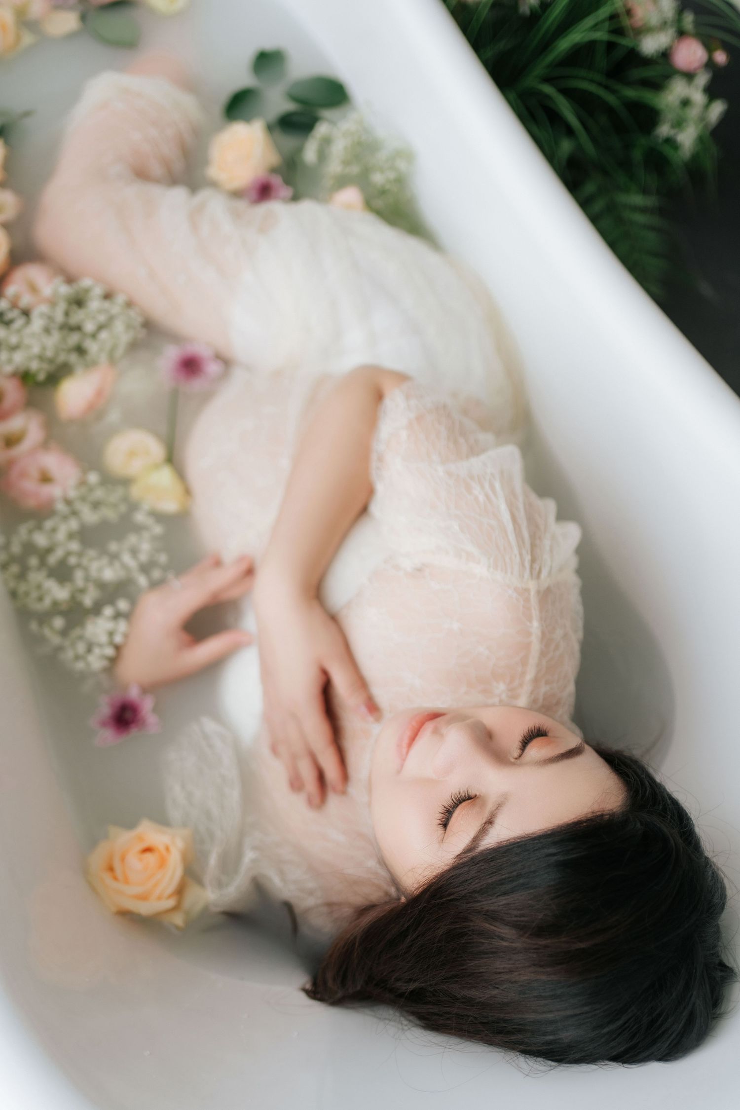 Woman in white lace robe relaxing in a floral milk bath surrounded by roses and greenery.身穿白色蕾絲睡袍的女子在玫瑰和綠植環繞的鮮花牛奶浴中放鬆身心。