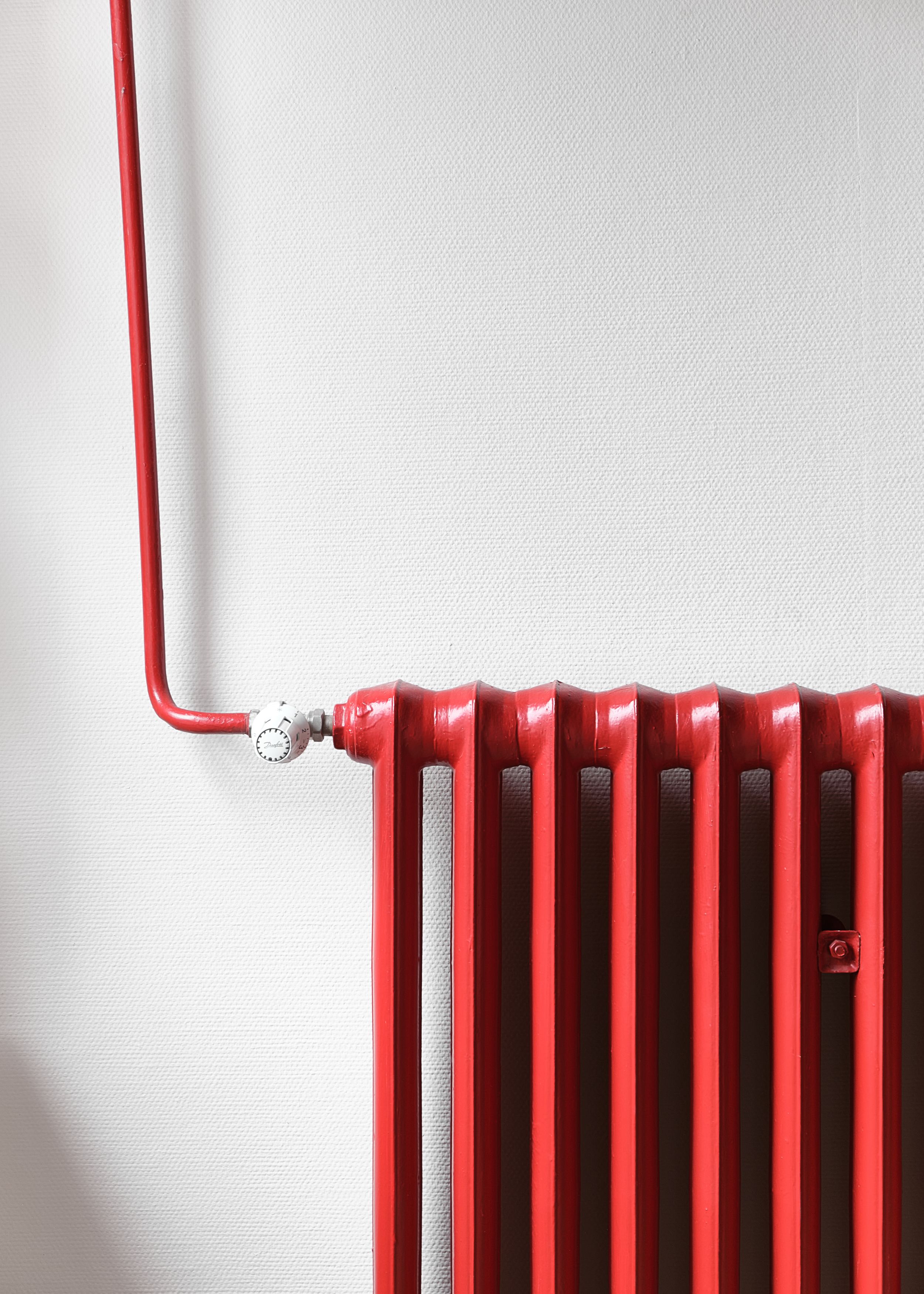 A bright red vintage radiator mounted on a white wall shows its distinctive ribbed design and exposed piping.