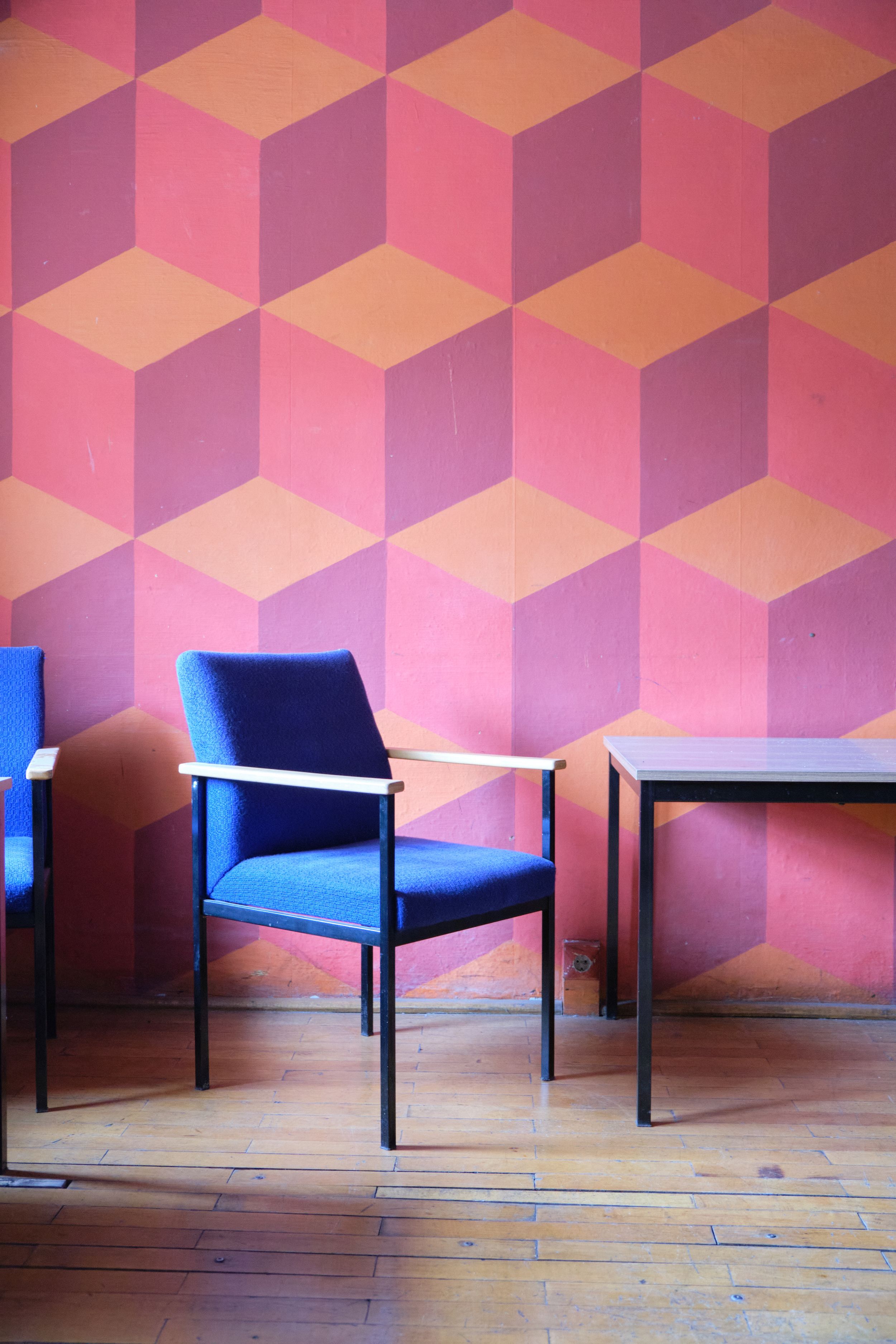 Modern blue chair against pink geometric wallpaper with cube pattern creates striking visual contrast on wooden floor.