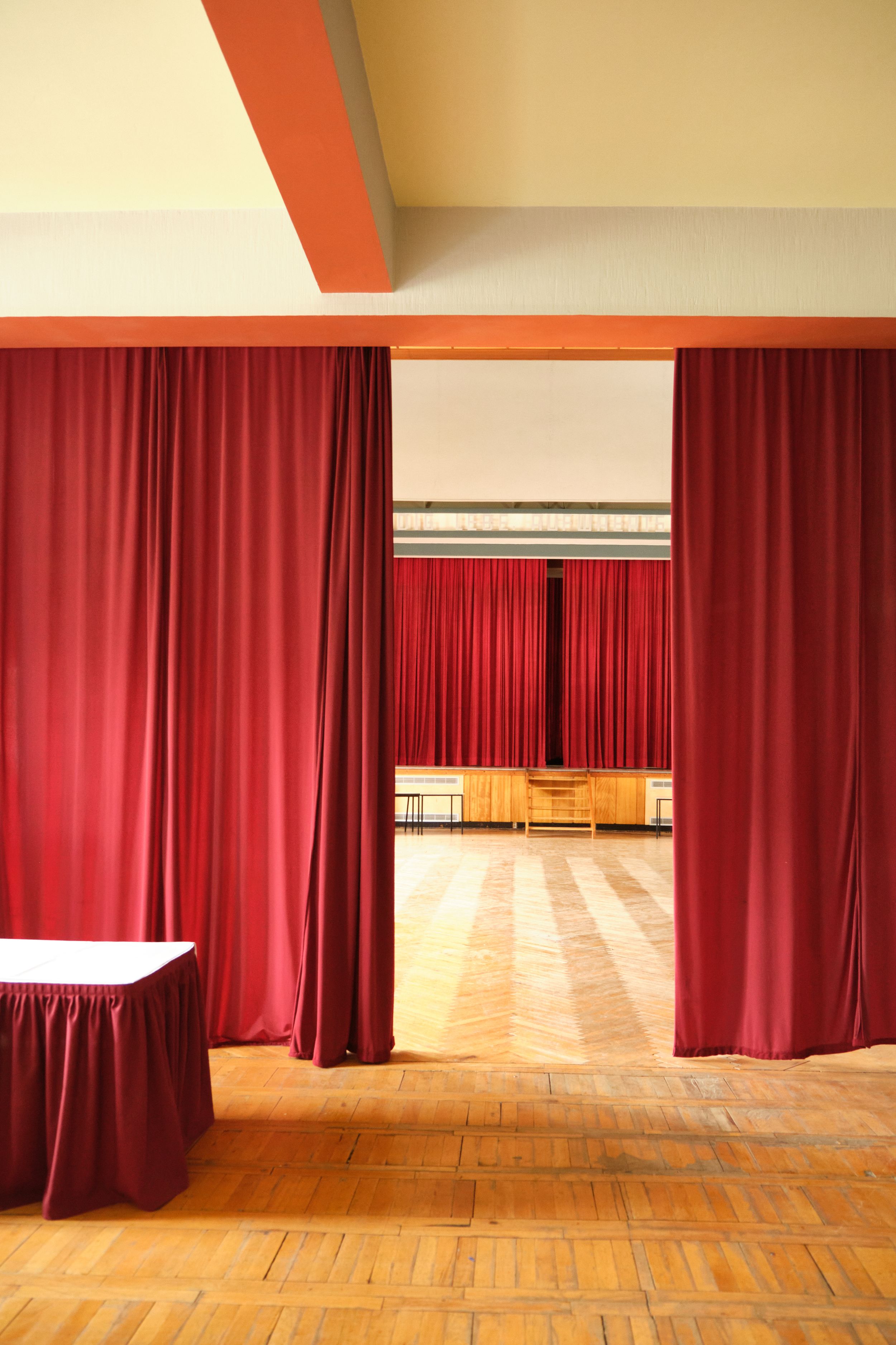 Red velvet curtains line a wooden floor stage with warm lighting and white walls in a performance space.