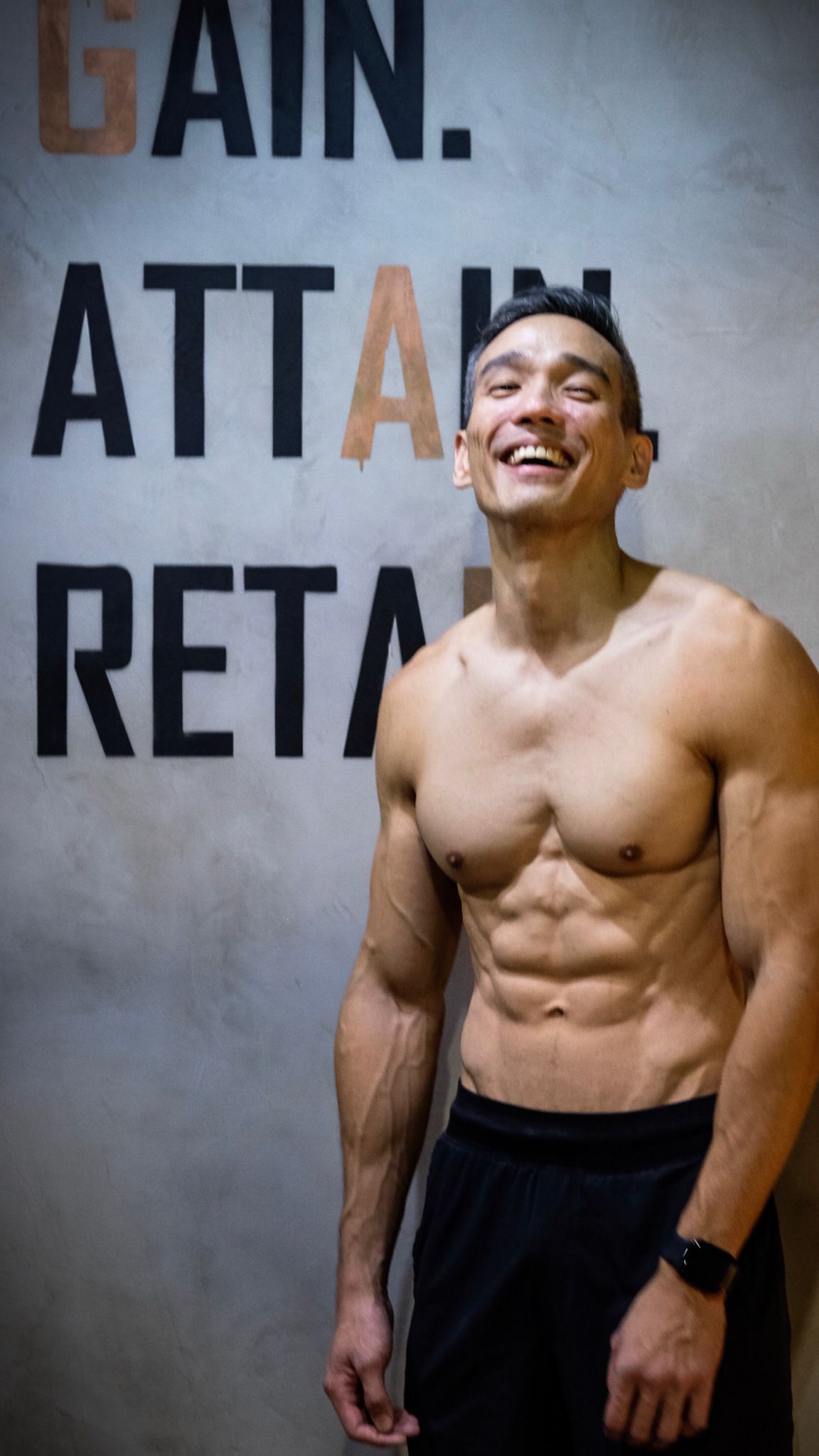 Fit shirtless man with six-pack abs smiling in front of motivational gym wall.