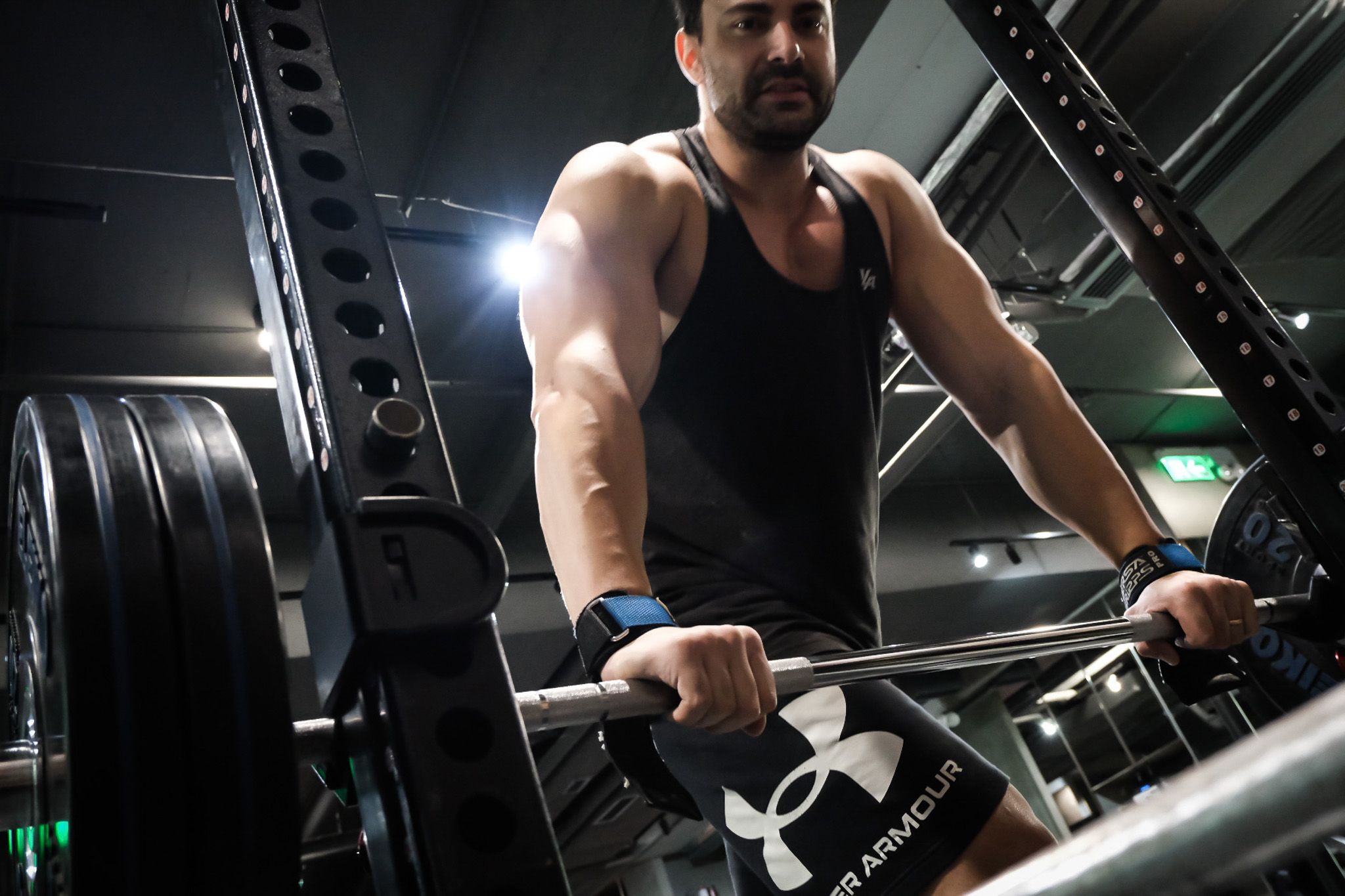 Muscular man in Under Armour gear performing barbell exercise in dark dramatic gym.