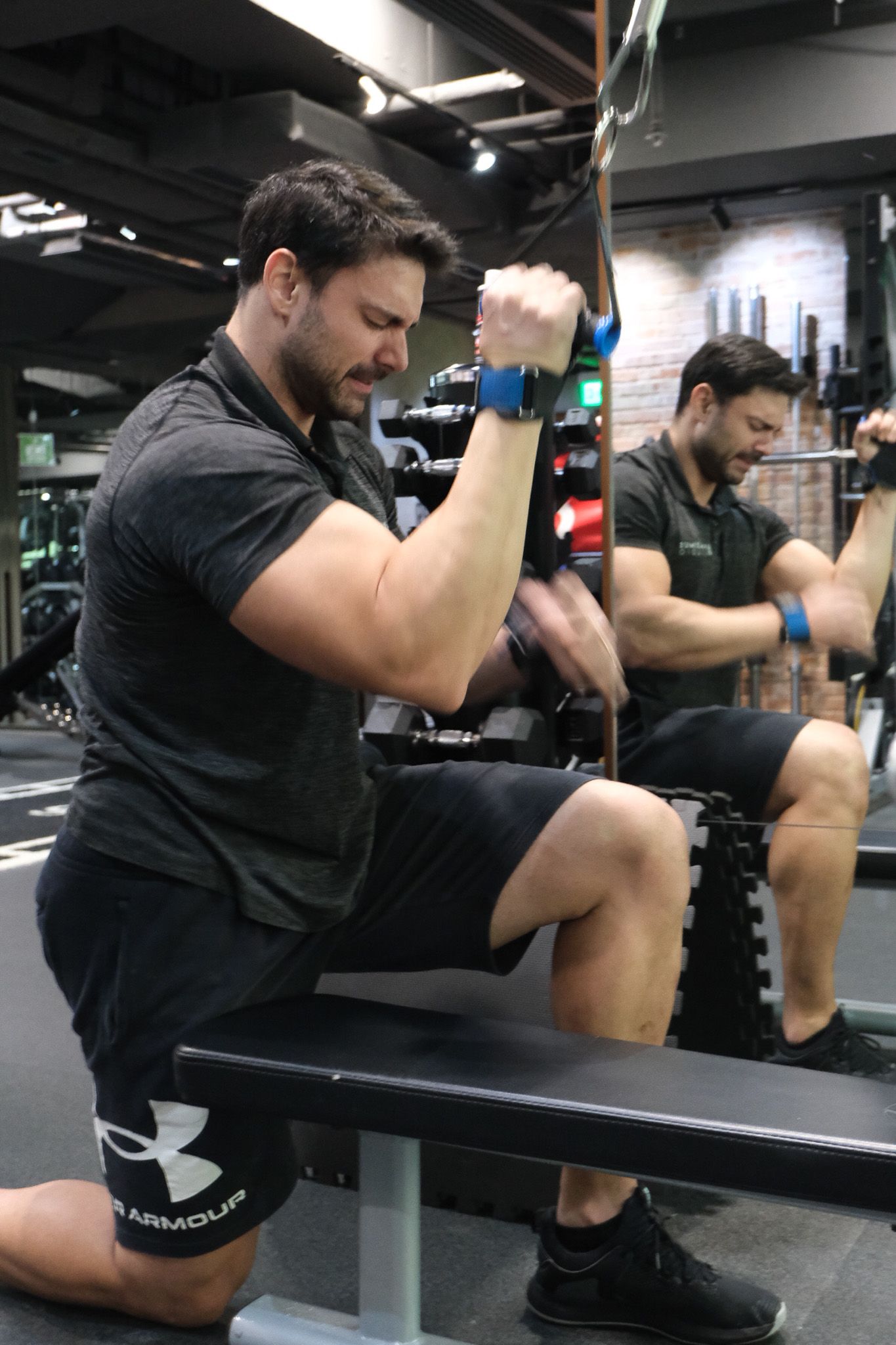 Muscular man in Under Armour shorts performing cable curl exercise on gym bench.