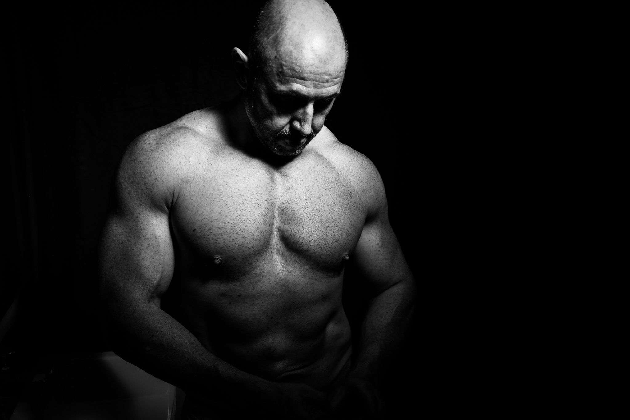 Black and white portrait of muscular bald man with defined physique on dark background.