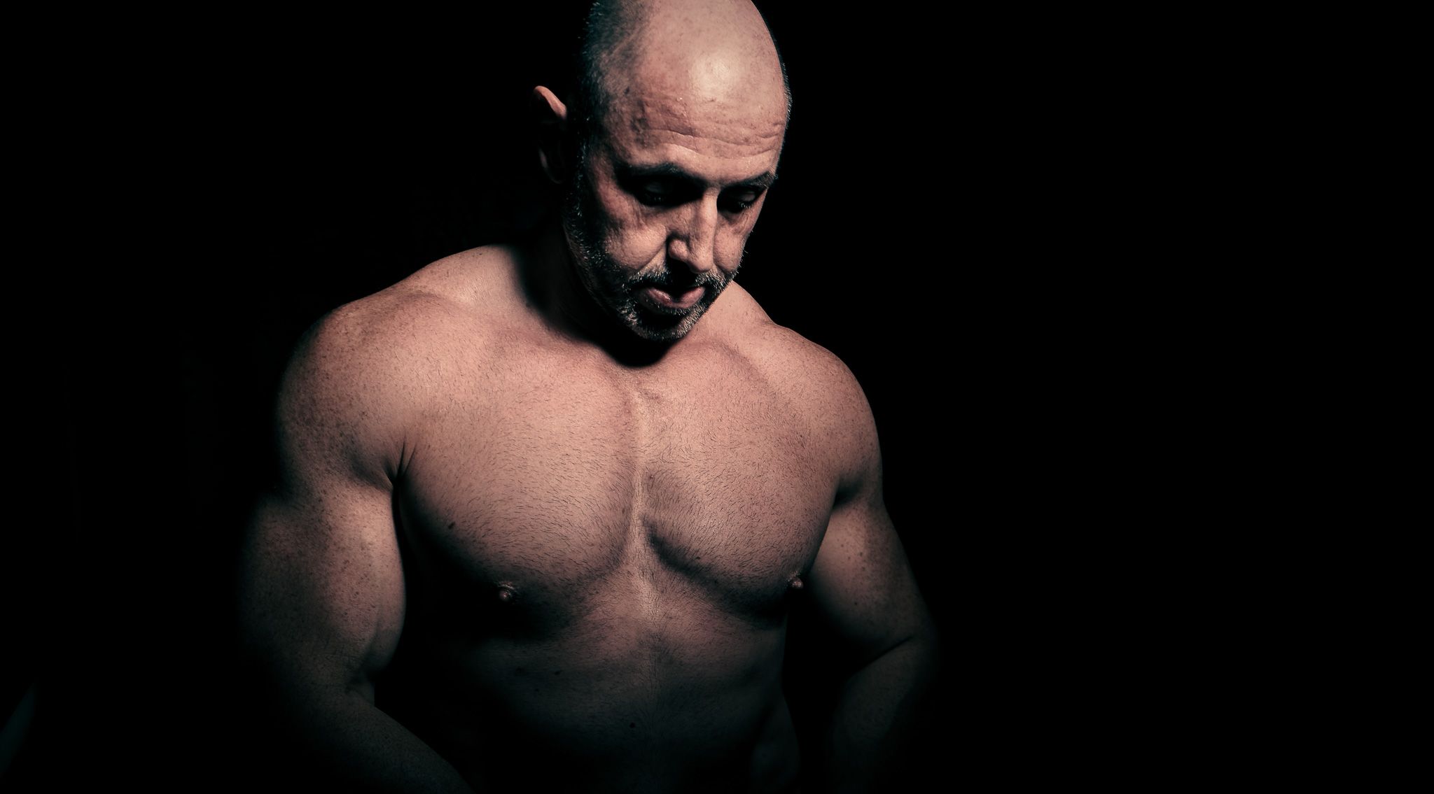 Dramatic low-key portrait of muscular bald man with defined chest in dark setting.
