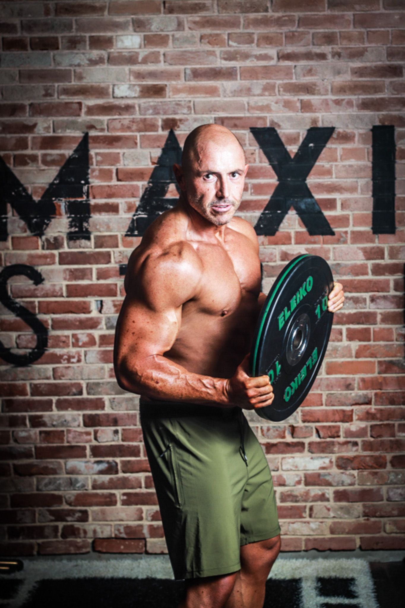 Muscular bald bodybuilder holding a weight plate in front of a brick wall with MAX text.