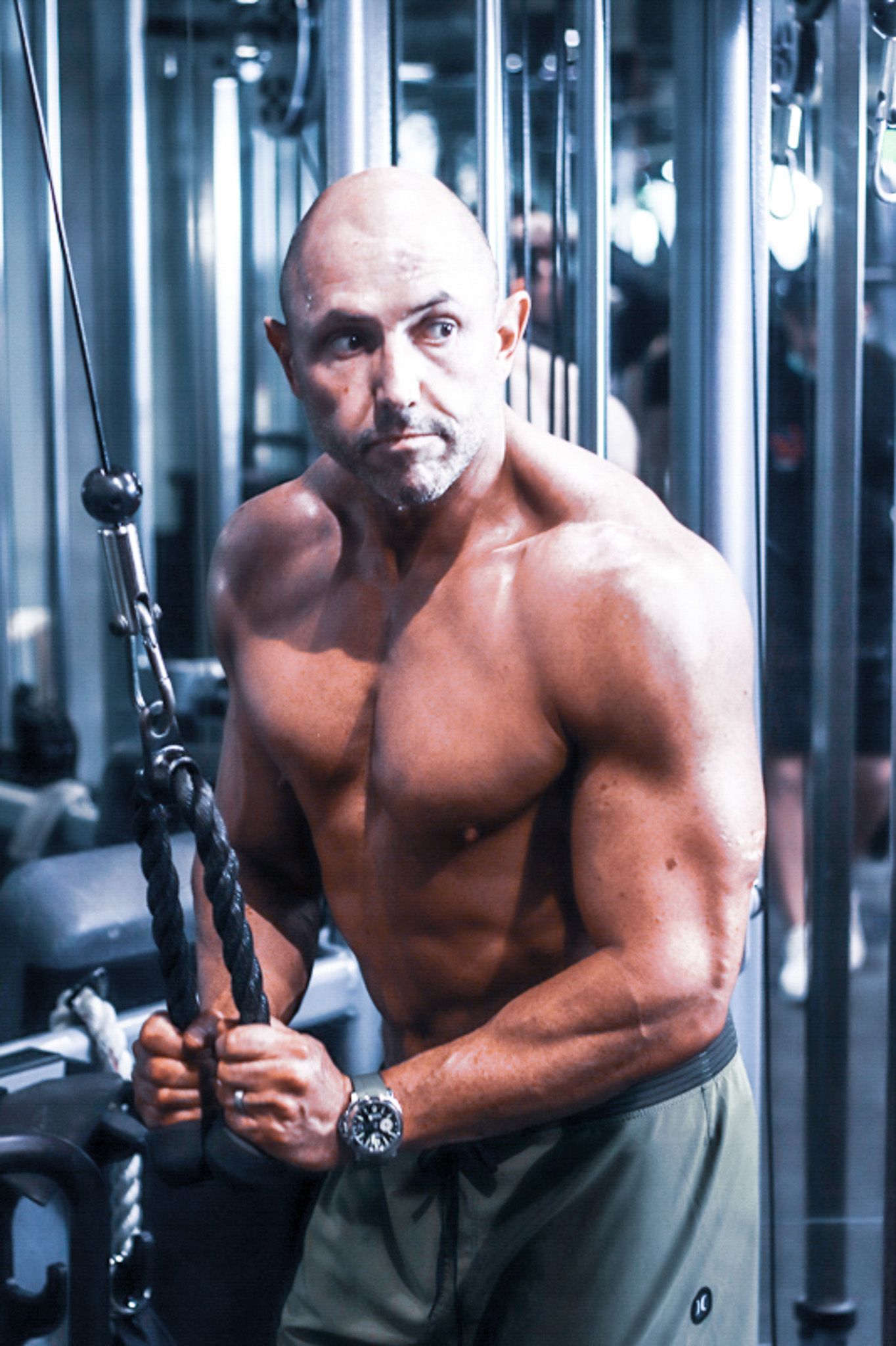 Muscular bald bodybuilder posing with cable rope attachment in a gym mirror setting.