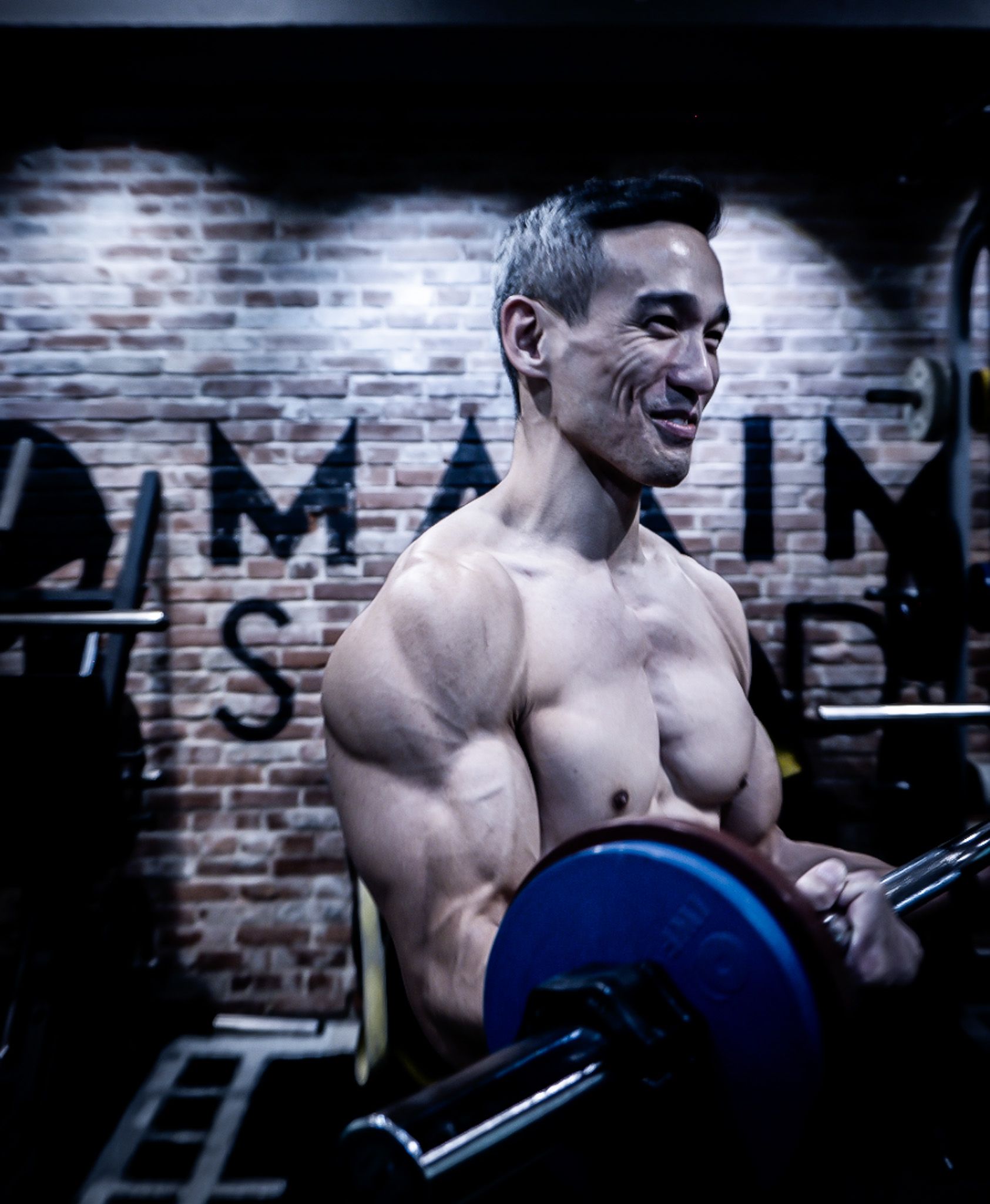 Buff Asian man smiling while lifting barbell in gym with exposed brick wall background.