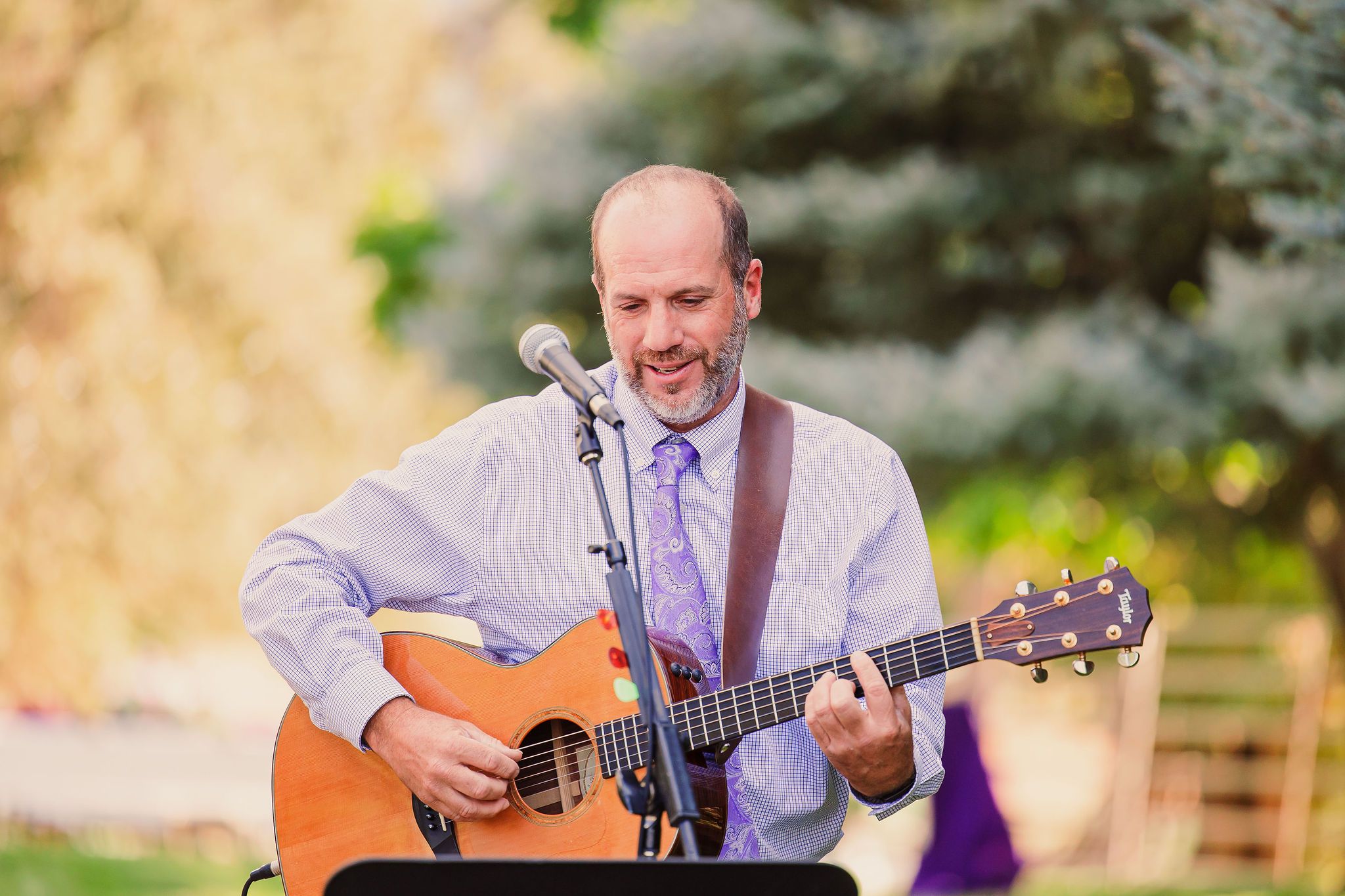 A musician performs outdoors with an acoustic guitar while wearing a purple dress shirt and tie.