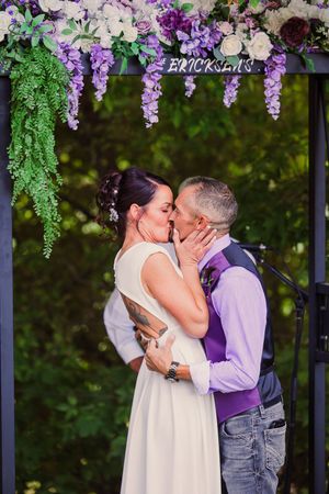 A couple shares a romantic kiss under purple wisteria flowers during an outdoor wedding ceremony.