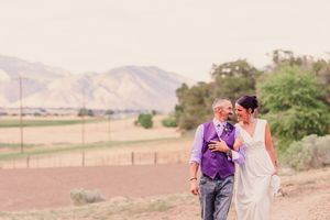A couple in purple and white outfits embrace while walking through a scenic desert landscape at sunset.