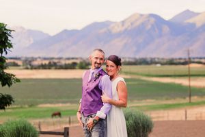 Couple embraces in golden field with majestic mountain backdrop during sunset photoshoot.