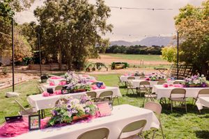 Outdoor wedding reception setup with white tables, pink decor and string lights under trees at sunset in a garden setting.