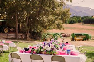 A rustic outdoor wedding reception setup with long tables decorated in pink linens under string lights and trees.