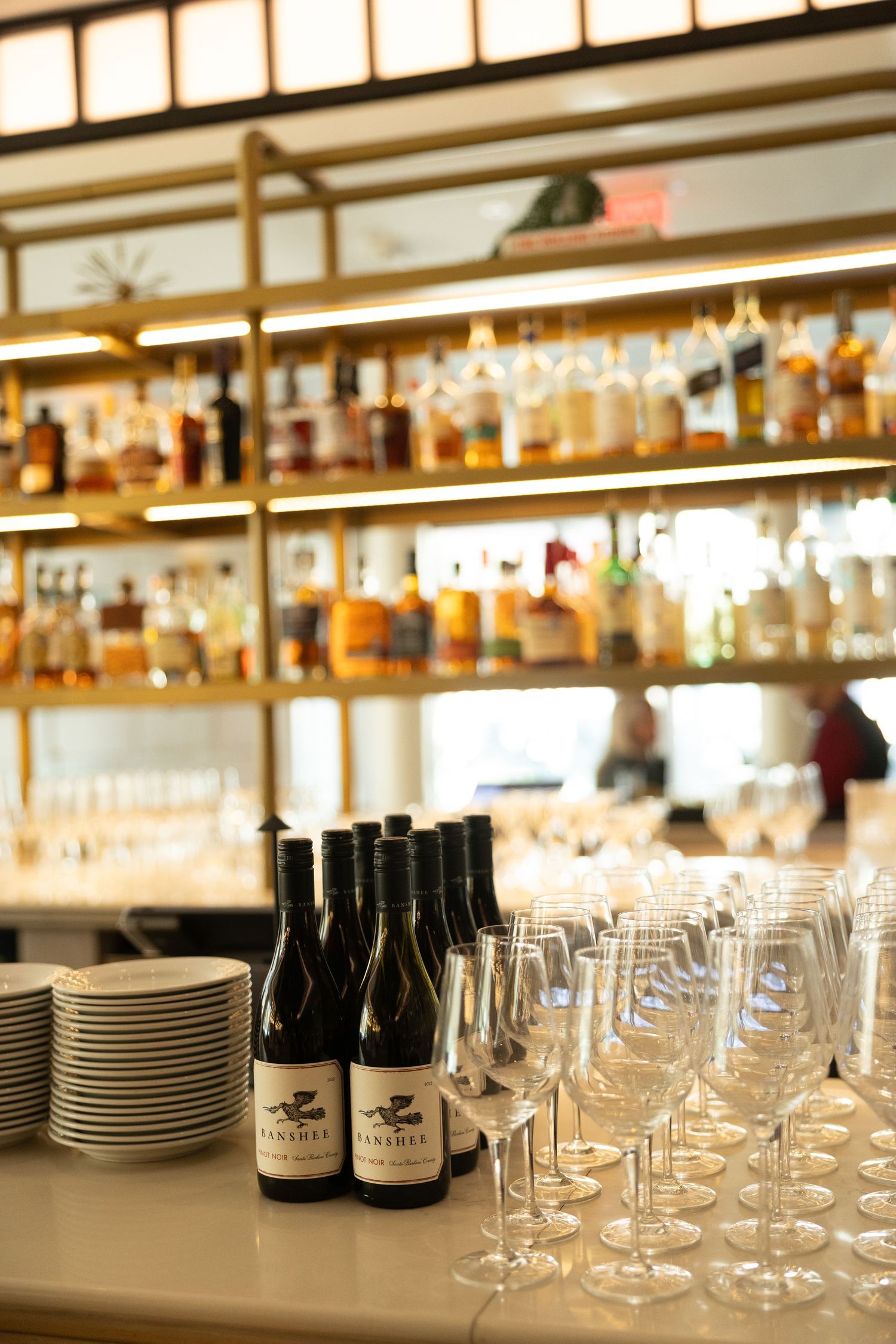 Wine glasses and plates arranged on a bar counter with bottles and warm lighting in an elegant restaurant setting.