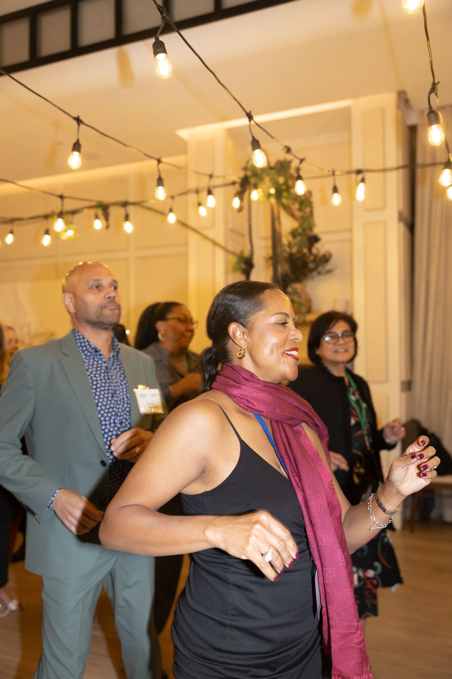 People dancing and celebrating at an indoor event with string lights and white walls.