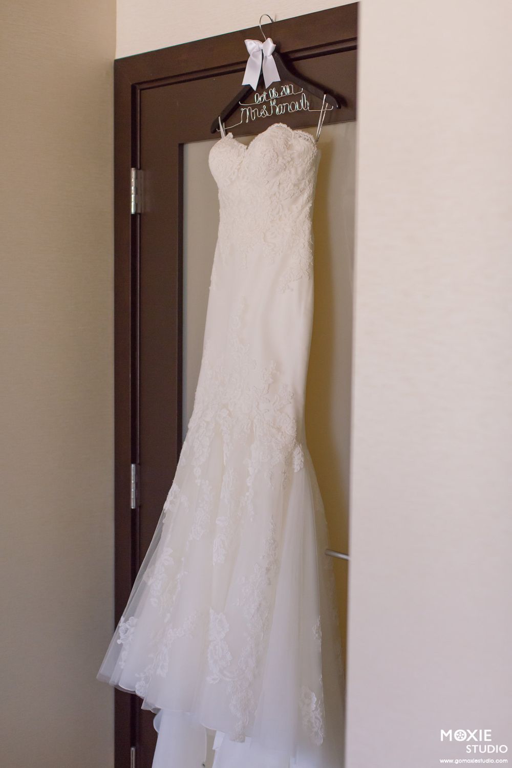 A white lace wedding dress hangs on a custom wooden hanger against a brown door.