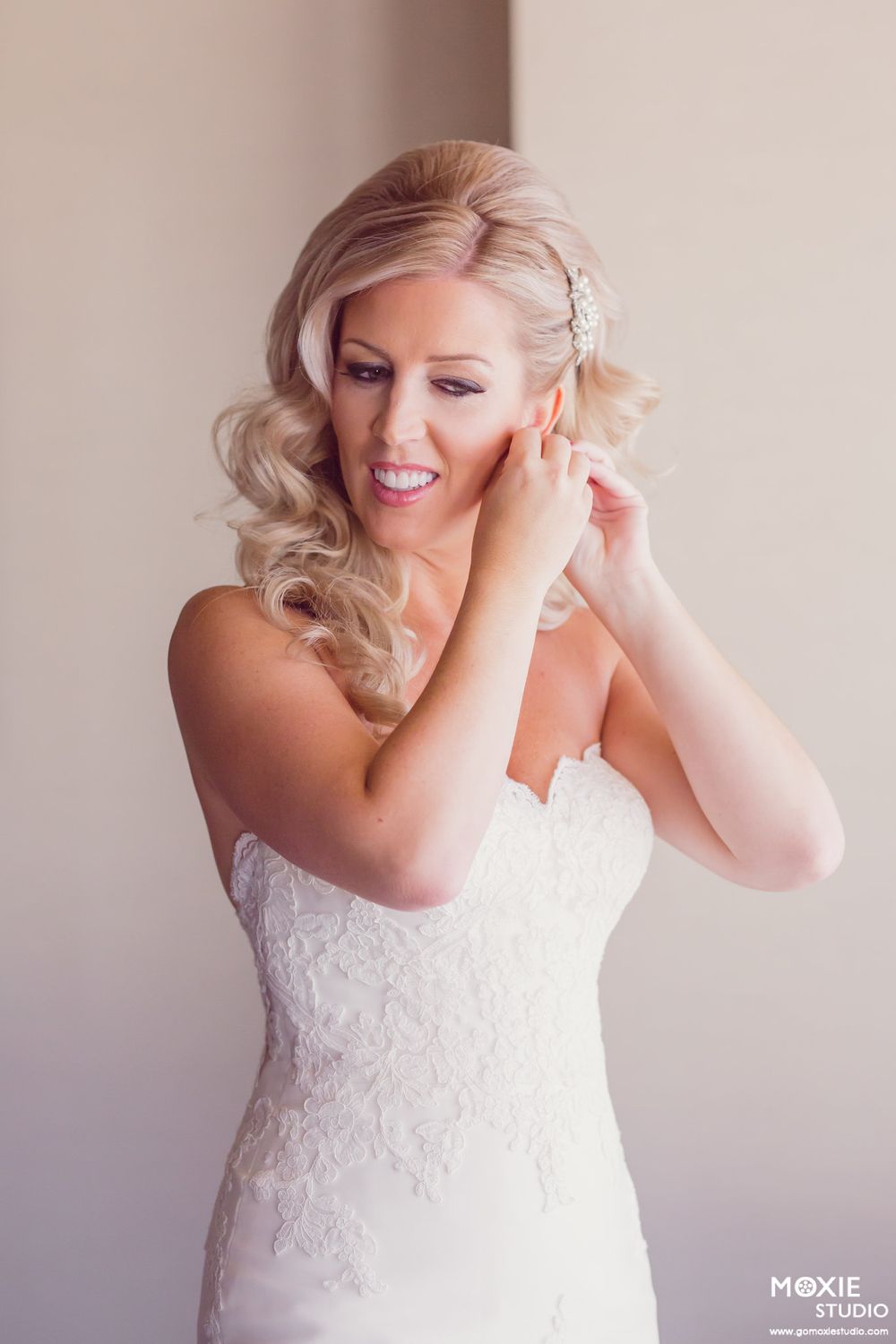 A bride in a white lace dress adjusts her earrings while getting ready in soft natural light.