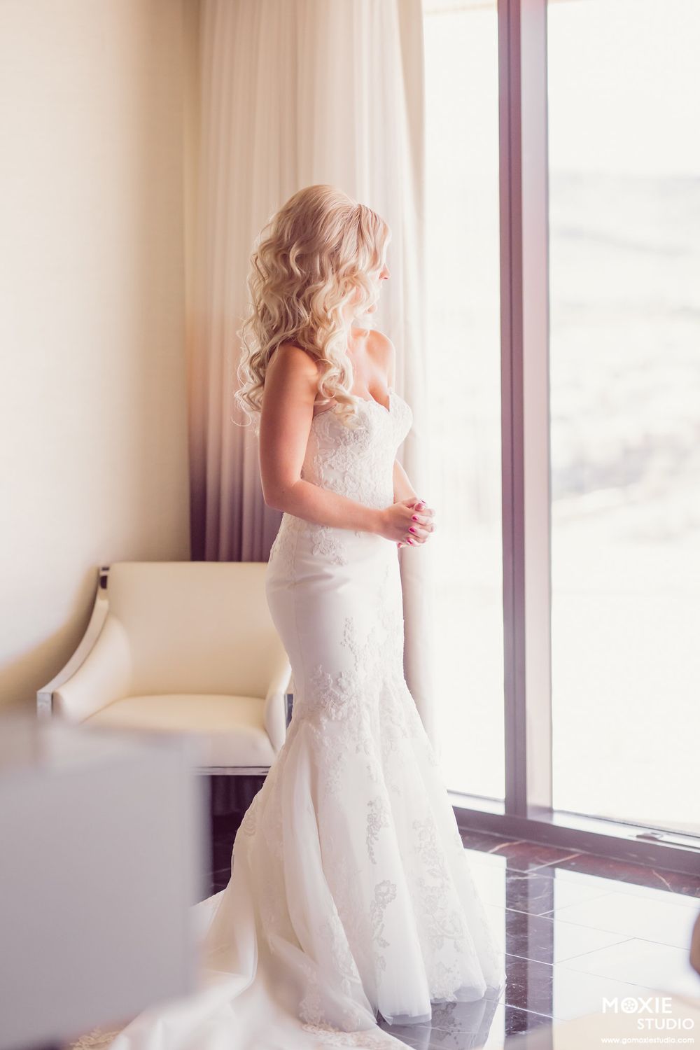 Elegant bride in fitted lace wedding gown stands by window in soft natural light.
