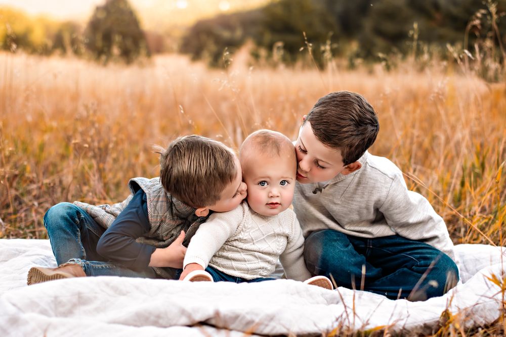 Baby sitting in field at Elk Meadow park being kissed by his two older brothers