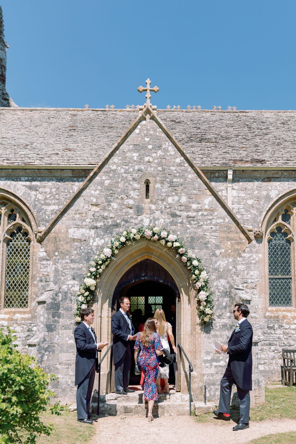 Wedding party gathers outside a historic stone church with gothic windows and floral arch decorations.