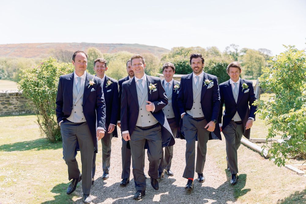 Groomsmen in matching navy suits and gray pants walking together on a sunny hillside path.