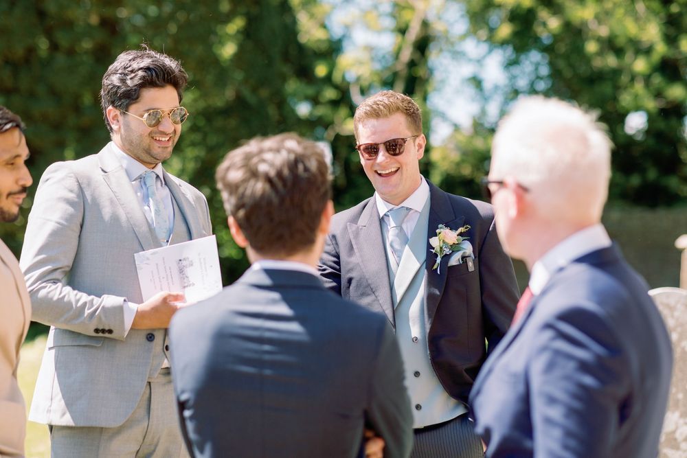 Groomsmen in light suits share a laugh during an outdoor wedding celebration on a sunny day.
